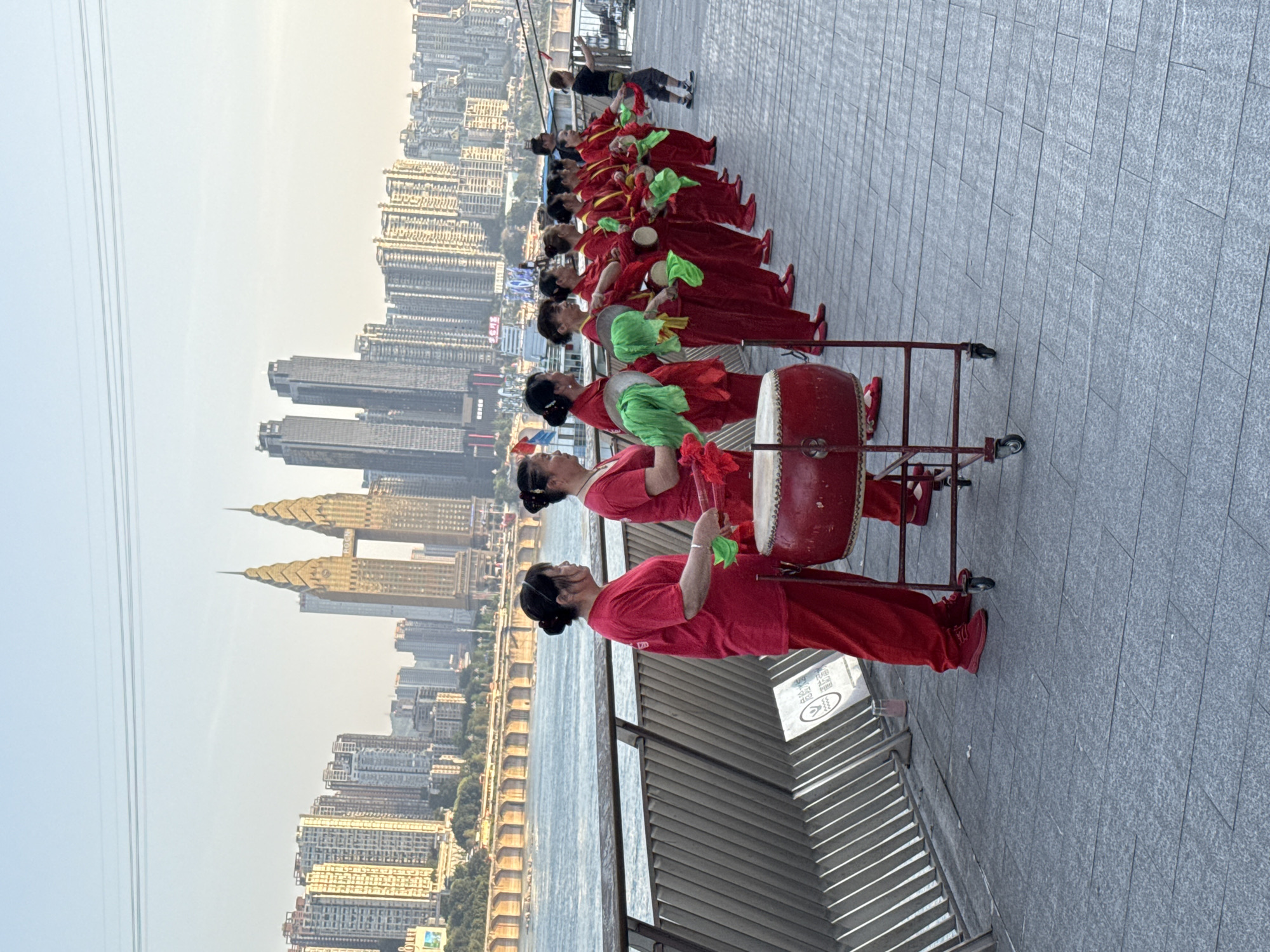A long line of women in red outfits, performing on a paved promenade along a river. Some women are holding a red drum on a wheeled cart. Many of the women hold green cloth accessories. Behind the group of women is a gray metal railing, the river, and a dense cityscape, including a distinct gold-colored twin-tower structure. The location appears to be Yuzhong District, China. A few people not in red outfits walk on the promenade behind the women performers.