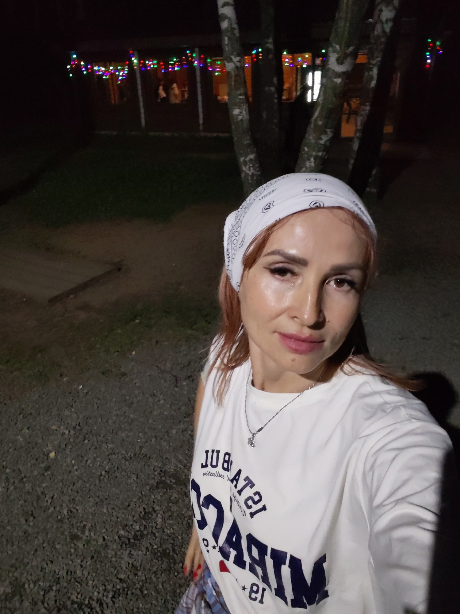 A mid-shot, waist-up, selfie is taken at night outdoors. The subject is a Caucasian woman with shoulder-length reddish-brown hair, wearing a white bandana with a black paisley pattern on her head, a white t-shirt with dark blue lettering that appears to say "ISTANBUL MIRACLE", and a silver necklace with a small charm. She has light makeup on, with noticeable blush and eyeshadow. She is looking directly at the camera with a slight smile. Behind her, string lights in various colors are visible, strung across a building with an awning. Several trees with light-colored bark are also in the background, some of which have the string lights draped around them. The ground is covered with gravel. The scene appears to be in a public outdoor area, possibly a park or recreational space, located in Chaikovsky, Russia, given the context.