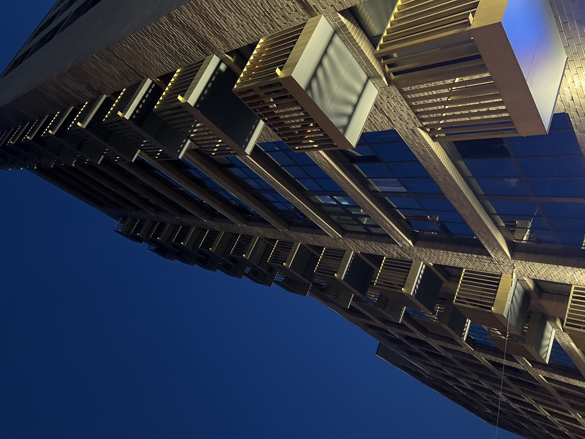 A low-angle, exterior shot captures the facade of a modern, multi-story brick building at dusk. The building features a repeating pattern of recessed balconies with light wood railings and white undersides. Integrated lighting casts a warm glow on the balconies and brickwork. Large, dark windows punctuate the facade. The deep blue of the twilight sky dominates the upper portion of the frame. The building is angled, with the camera looking up towards its upper floors. The location is identified as Unknown, Russia.