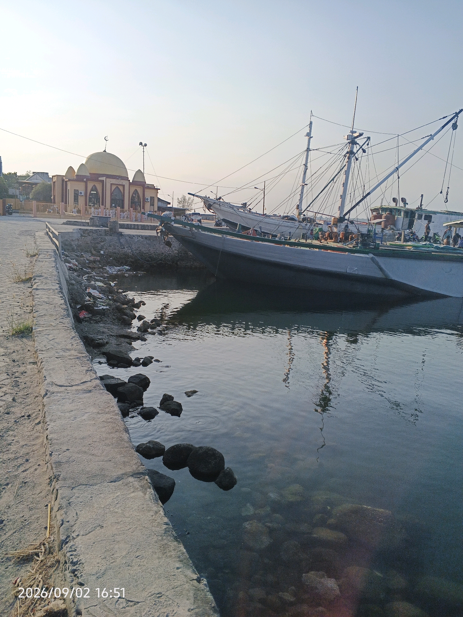 The image depicts a waterfront scene in Pare-Pare, Indonesia, featuring a mosque with a golden dome and several fishing boats docked along the shore. The mosque, located on the left side of the frame, is a prominent building with arched windows and a crescent moon atop its dome. In the foreground, a concrete walkway runs along the water's edge, leading towards the boats. The water is calm and reflects the sky and the boats. The shore is rocky and littered with some trash, suggesting a potentially unkempt area. The fishing boats are large vessels with masts and rigging, indicating their use for commercial fishing. The sky is clear and bright, suggesting it is daytime. The overall impression is a functional harbor area adjacent to a place of worship, with a mixture of natural and man-made elements. The timestamp "2026/09/02 16:51" is visible in the bottom left corner, indicating the date and time the photo was taken.