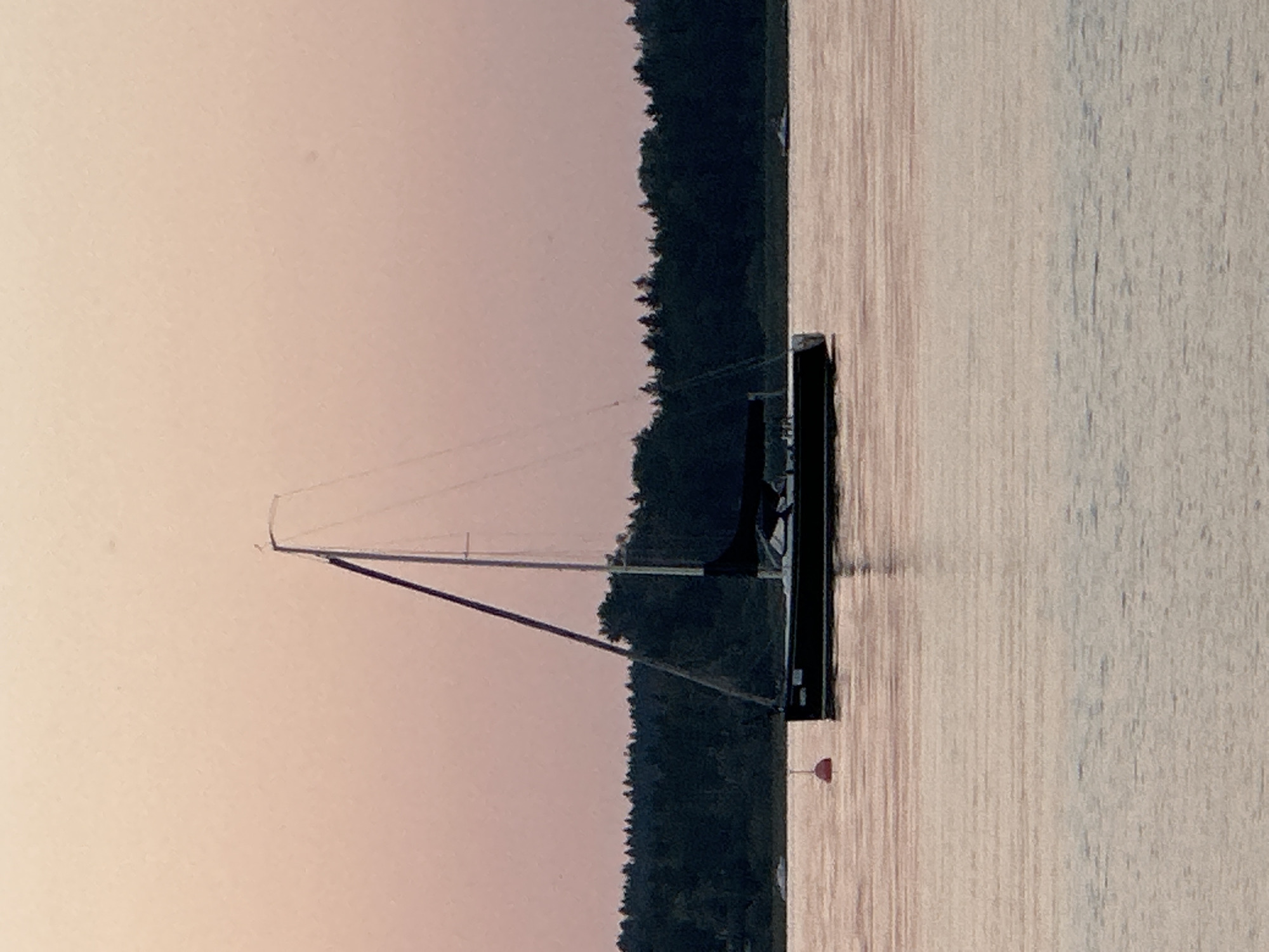 The image depicts a sailboat on a body of water, likely a lake or bay, at dusk. The sky is a soft, peachy-pink, transitioning to a lighter hue towards the horizon. A dark green treeline lines the background, suggesting a forested shore. The water is calm, reflecting the pastel colors of the sky and the silhouette of the boat.
The sailboat is dark-colored and has a single mast with rigging visible. A dark sail is partially furled. A small, red buoy is visible in the water to the left of the sailboat. In the distance, another small white boat can be seen near the treeline. There are no people visible in the image. The scene appears tranquil and serene.
While the image itself does not provide definitive clues to its location, the description requests a potential location in Prien am Chiemsee, Germany. Prien am Chiemsee is situated on the shore of Lake Chiemsee, a large Bavarian lake known for its scenic beauty and sailing opportunities. The presence of a sailboat, treelined shores, and calm waters aligns with the characteristics of Lake Chiemsee. However, without further contextual information or identifiable landmarks, this specific location remains speculative based on the provided image.