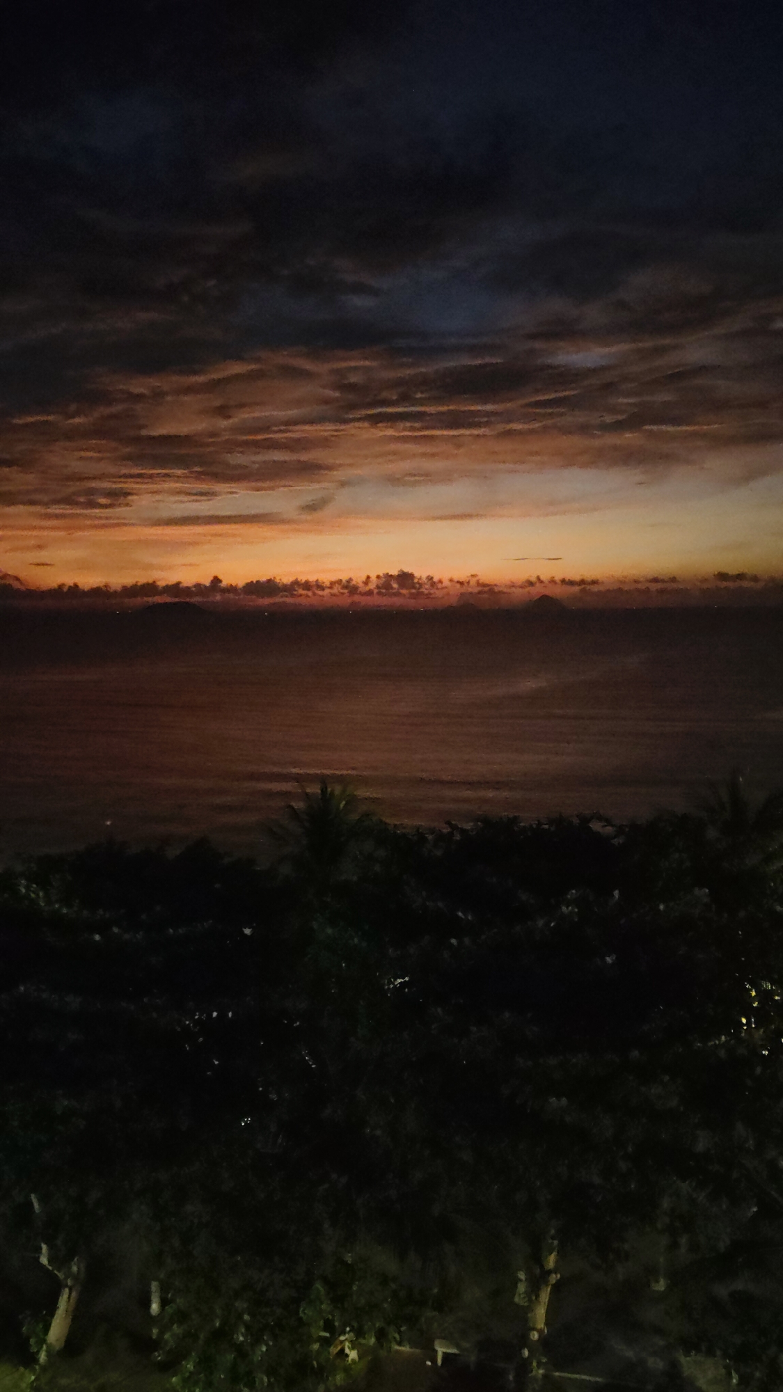The image shows a nighttime scene in or around Bac Nha Trang Ward, Vietnam. The view is elevated, looking out over a body of water towards the horizon. The sky is predominantly dark with clouds, exhibiting shades of orange near the horizon line, suggesting a sunset or sunrise. The water is dark, reflecting the colors of the sky. In the foreground, there are dense, dark trees and vegetation, obscuring the lower part of the image. The overall impression is of a dimly lit landscape at dusk or dawn.