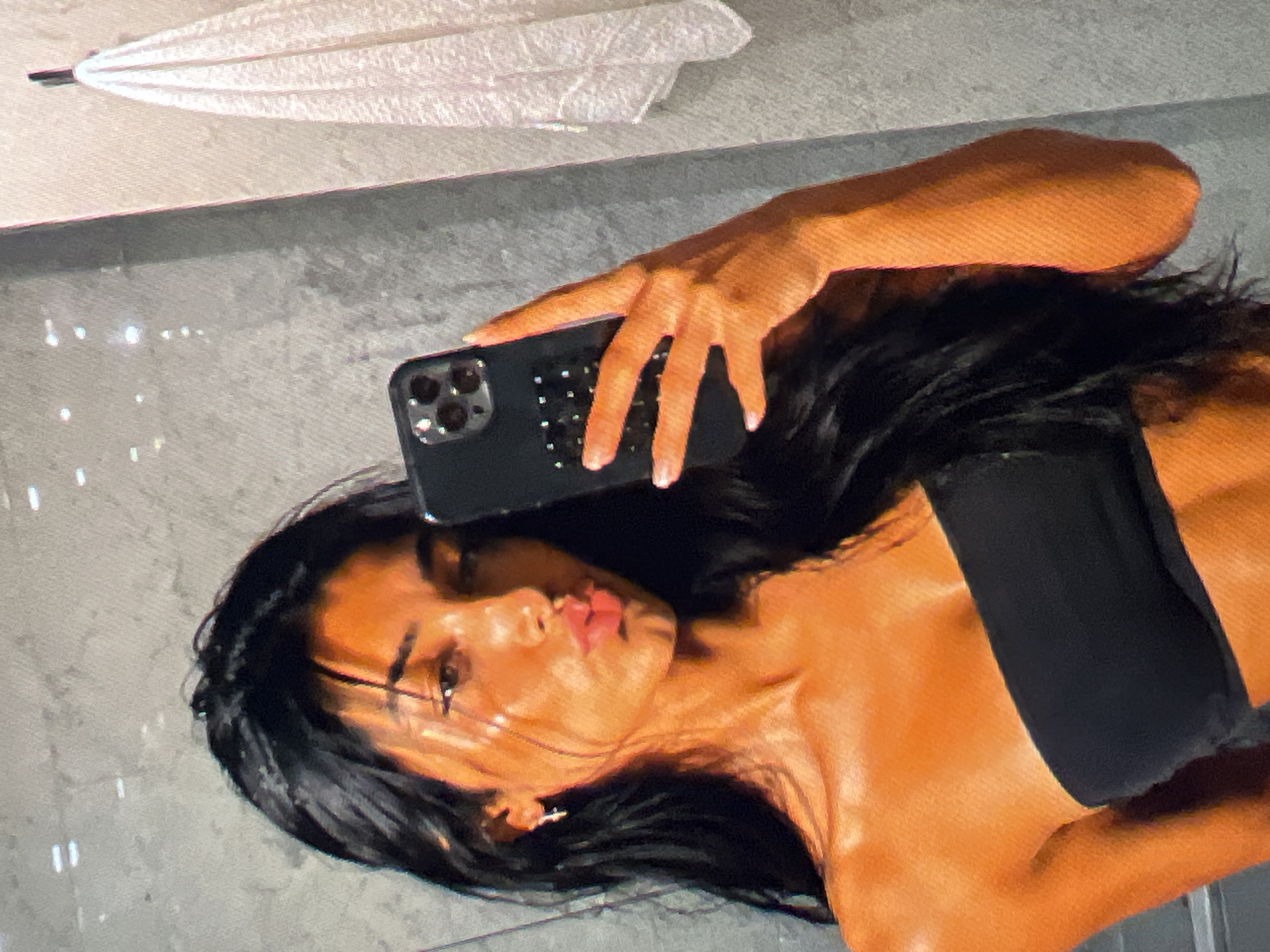 An individual with long, dark hair and tan skin is taking a selfie in a bathroom. They are wearing a black bandeau top. Their mouth is puckered as if blowing a kiss. They are holding a dark-colored smartphone in their right hand, which is angled to capture the reflection in the mirror. The background of the mirror shows a textured gray wall, and to the right, a white towel is hanging on a hook. The lighting in the bathroom casts a warm glow on the individual's skin, highlighting their features and musculature.