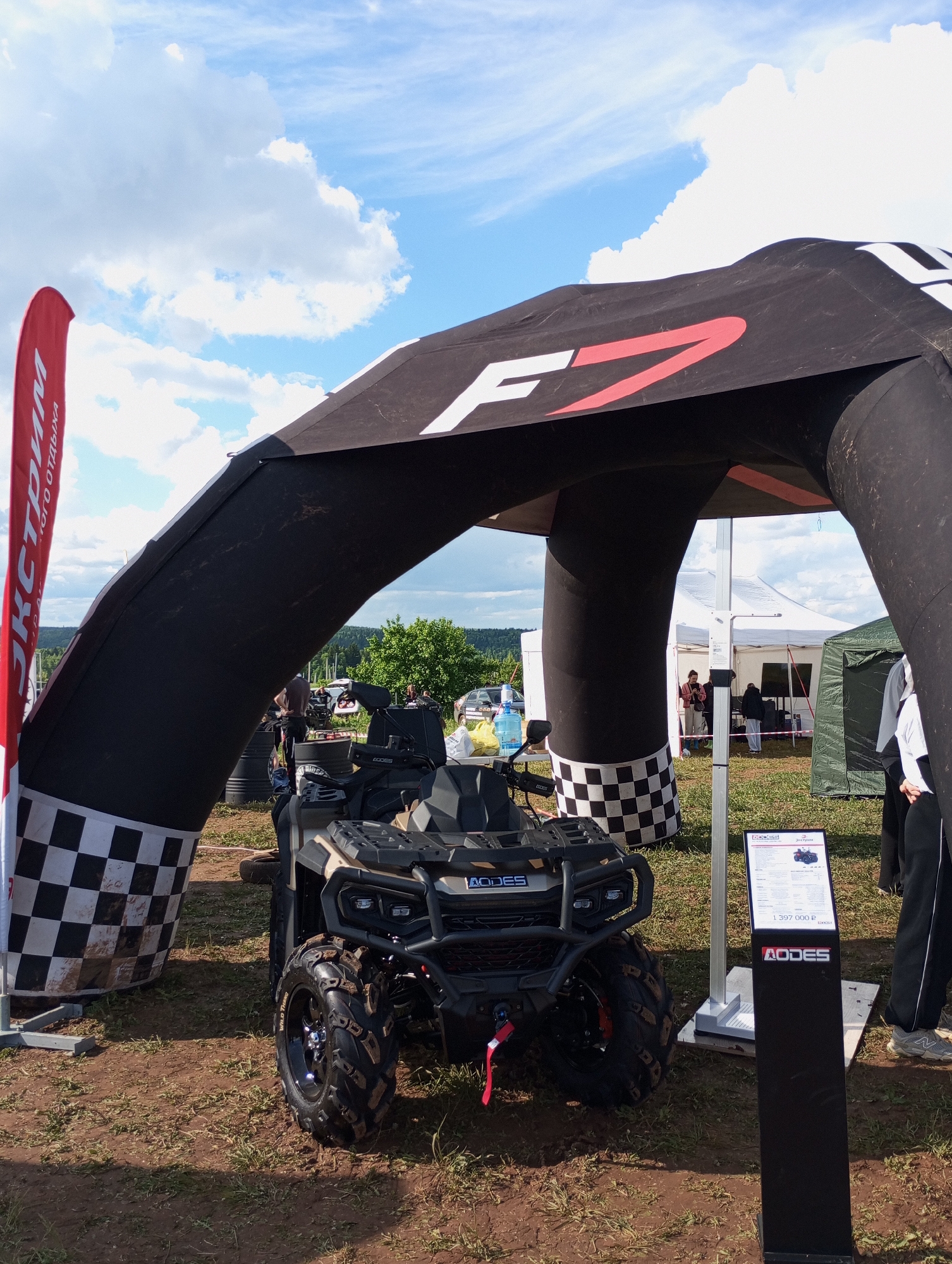 The image shows an AODES all-terrain vehicle (ATV) displayed outdoors in Izhevsk, Russia. The ATV is positioned under a black inflatable archway featuring checkered flags and the letter "F" in red and white. A red banner with Cyrillic text is adjacent to the archway. To the right of the ATV, a black informational display stand with the AODES logo and pricing details is visible. In the background, tents, vehicles, and people are visible, suggesting an event or exhibition. The environment appears to be a grassy field under a partly cloudy sky.