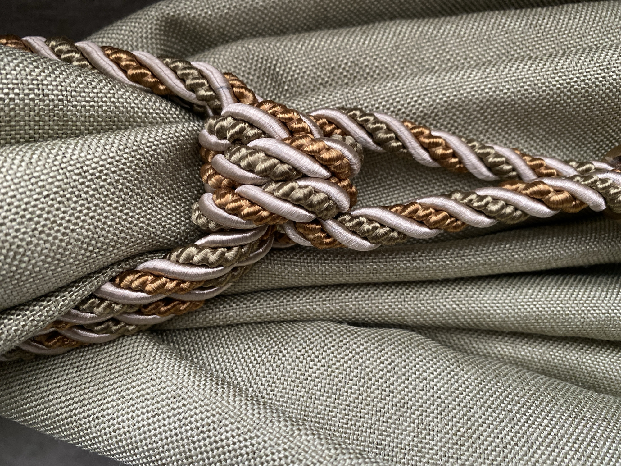 The image depicts a curtain tieback draped over a folded curtain. The curtain appears to be made of a textured, light-colored fabric, possibly linen or a similar material with a visible weave. The tieback is a decorative cord with a complex knot. The cord is made of twisted strands in three colors: a light beige, a golden brown, and a light green. The knot is centrally located, pulling the curtain fabric together in soft folds. The background is blurred, suggesting a shallow depth of field. The lighting is soft and diffused, illuminating the textures of the fabric and cord.
The location is not known.