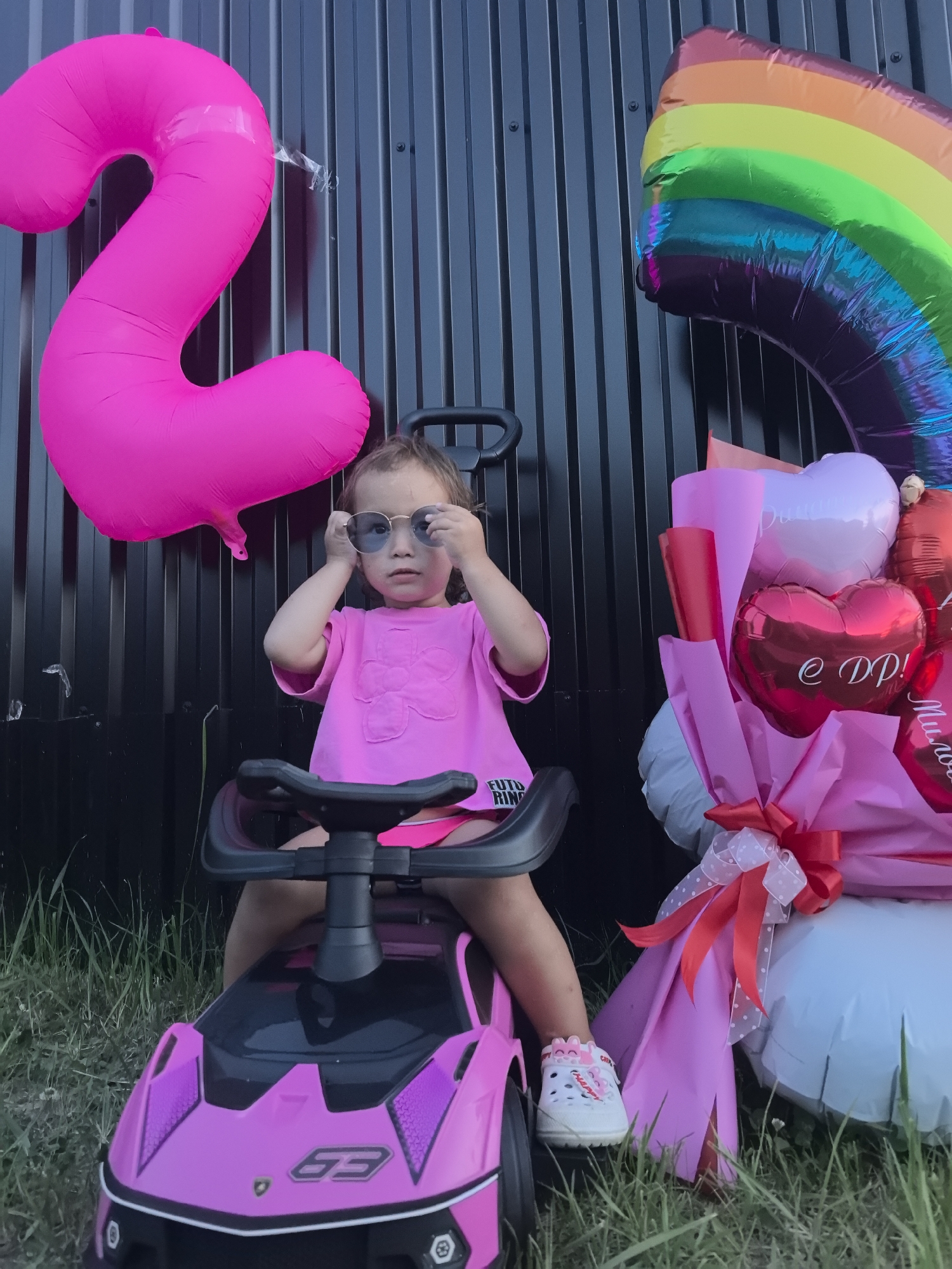 A young Caucasian girl with light brown hair is sitting in a pink toy car. She is wearing a pink t-shirt and pink shorts, and white crocs with bunny ears. She is wearing sunglasses and is looking at the camera. Behind her is a black corrugated metal fence. To her left, a large pink number "2" balloon is attached to the fence. To her right, a bouquet of pink and red balloons is visible, along with a rainbow-colored balloon. The ground is covered in green grass. The scene appears to be an outdoor celebration, possibly a birthday party. The location is Chaikovsky, Russia, as indicated by the context.