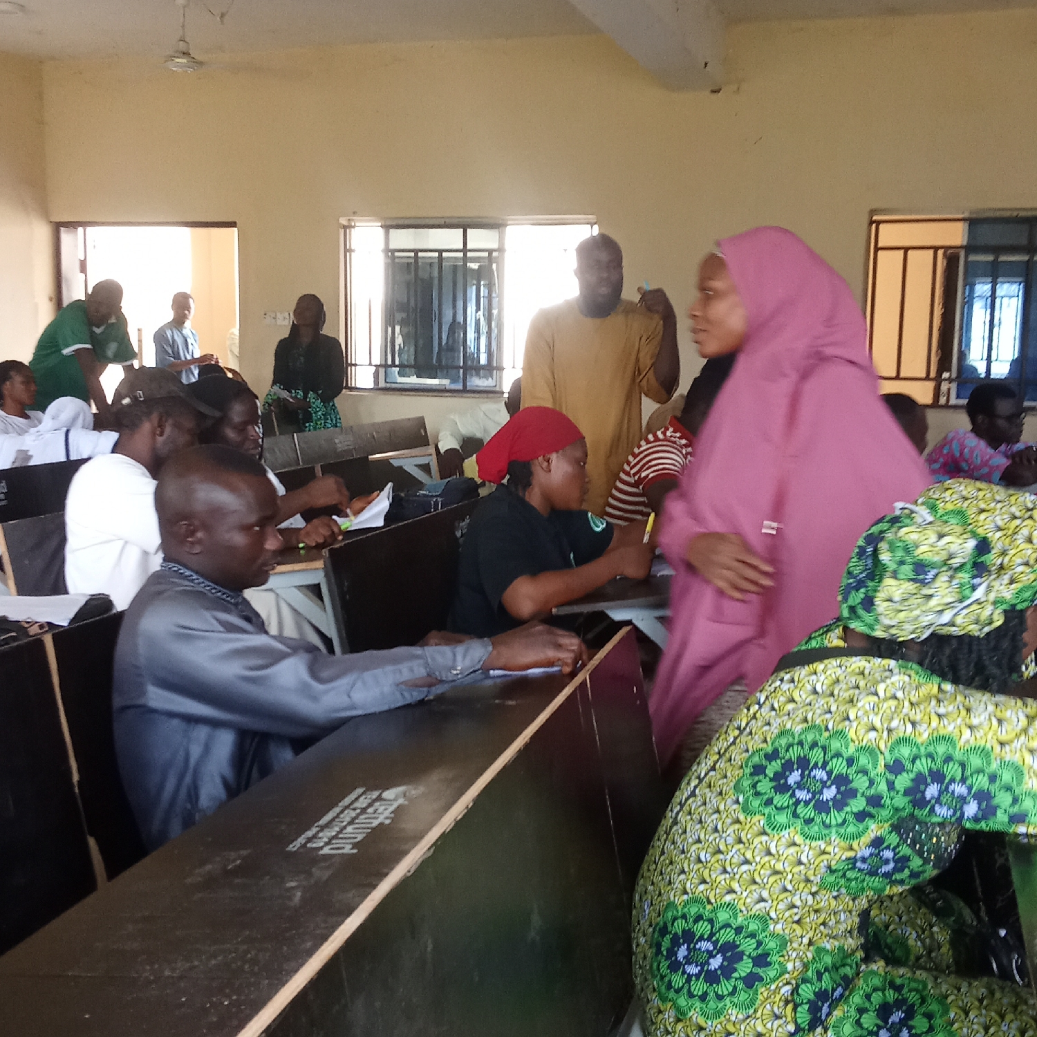 The image depicts an indoor gathering of approximately 15-20 adults, predominantly Black individuals, within what appears to be a classroom or training room in Akwanga, Nigeria. The room is brightly lit by natural daylight, suggesting it is daytime. Light-colored walls and a ceiling fan are visible, along with multiple windows fitted with dark metal bars.
In the foreground, a woman wearing a prominent pink hijab is seen in motion, walking through an aisle. To her right, another woman is visible from behind, dressed in a vibrant green, yellow, and black patterned fabric, likely a traditional Ankara print. A man in a grey traditional outfit (often called a 'senator' suit in Nigeria) is seated at a dark wooden desk in the mid-ground, leaning forward, seemingly writing or focusing on materials. The word "eFund" is visibly printed on the surface of this desk.
Throughout the room, other attendees are seated at similar desks, some appearing to be engaged with papers or notebooks, while others are standing or interacting. In the mid-ground, a man in a tan, short-sleeved top stands, seemingly conversing with the woman in pink. In the background, near the windows, another man in a green t-shirt and a woman in a dark patterned dress are also present. The overall scene suggests a busy workshop, lecture, or meeting in progress, or during a transitional moment. The attire of several individuals provides cultural context consistent with the given location.