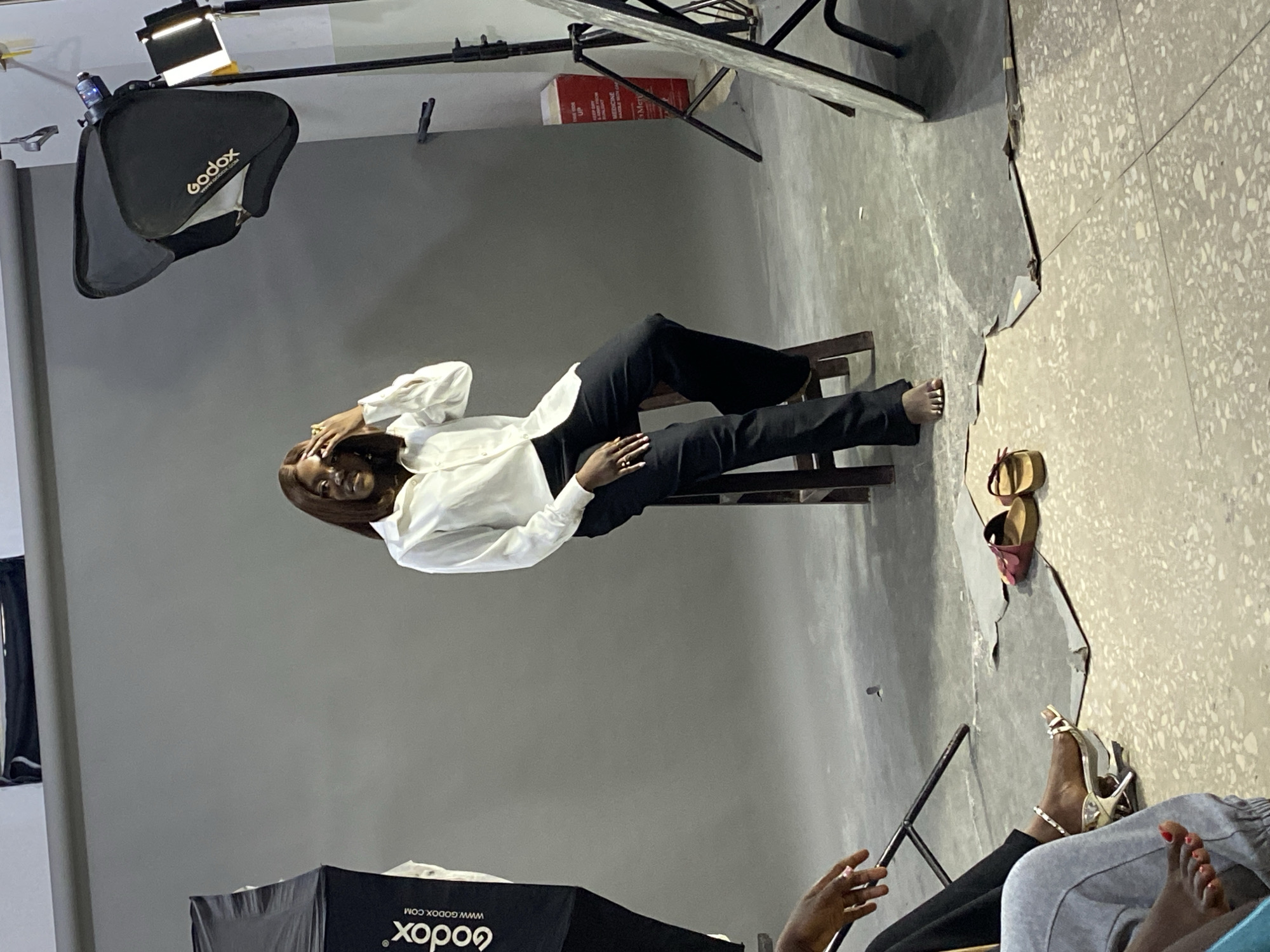 The scene is a photography studio, possibly located in Jos, Nigeria, given the information provided. A woman is seated on a wooden chair in front of a grey backdrop. She is wearing a white shirt and black pants, and her hair is styled in a short brown bob. She is posed with her hand near her head. Photography lighting equipment, including a Godox softbox, is visible around her. Other objects in the scene include shoes near the corner of the backdrop and other seated people, feet are seen, possibly members of the photography crew.