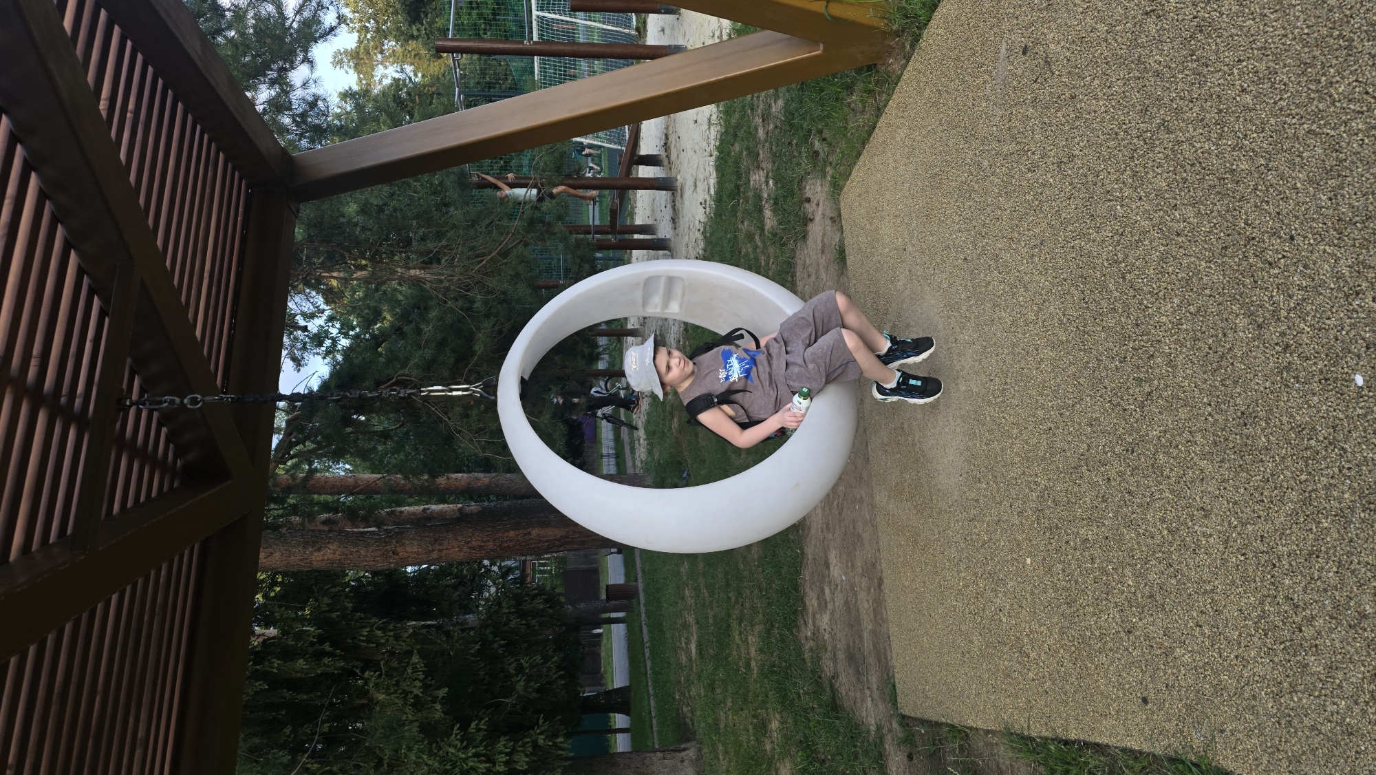 A young boy wearing a grey hat, a grey t-shirt with a blue graphic, grey shorts, and black and blue sneakers is sitting in a white, circular swing suspended from a wooden pergola. He is holding a green and white bottle. The swing is situated on a sandy area with a speckled yellow surface underneath. In the background, there are trees, a playground with metal bars, and other children. The location is Il'inskiy, Russia.