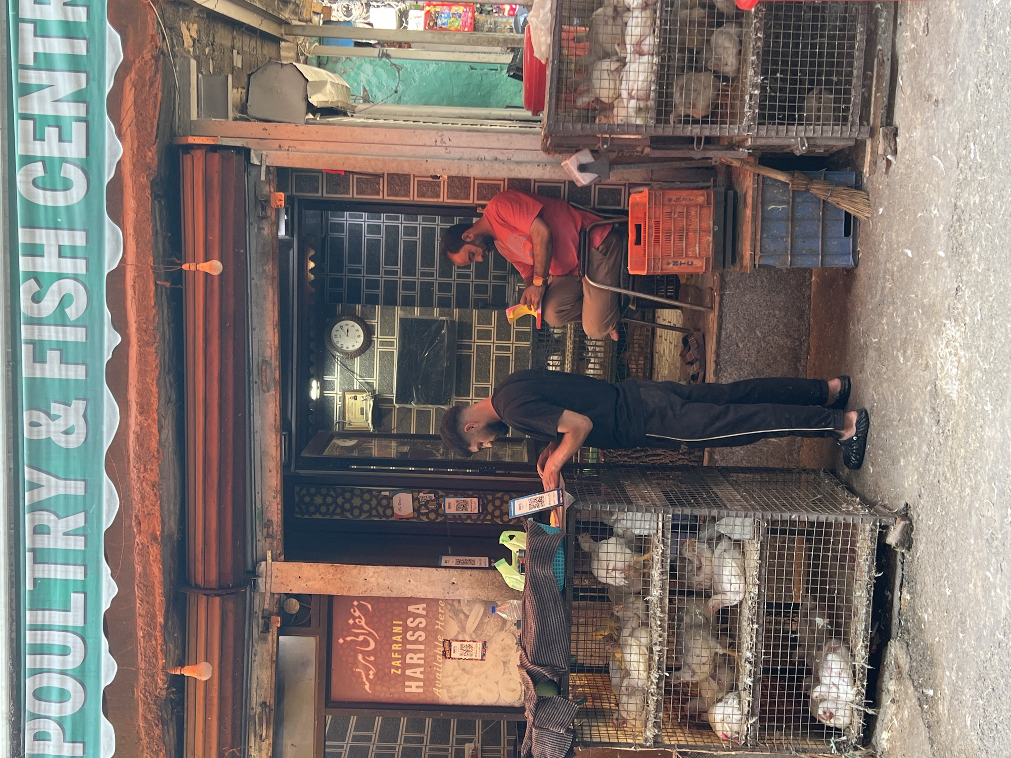 This image captures a street scene in Srinagar, India, outside a shop labeled "POULTRY & FISH CENTRE." Two men are present. In the foreground, a man wearing a black t-shirt and black pants stands, facing a laptop and engaging with it, possibly a point-of-sale transaction. In front of him are several cages containing live chickens. To his right, a man in a red t-shirt and brown pants sits on a chair, looking at a yellow object in his hands. The shop's entrance is visible behind them, with a sign in Urdu and English advertising "ZAFFRANI HARISSA Available Here." The environment appears to be an outdoor market setting, with various crates and a broom visible on the ground. The overall impression is of a bustling, functional marketplace.