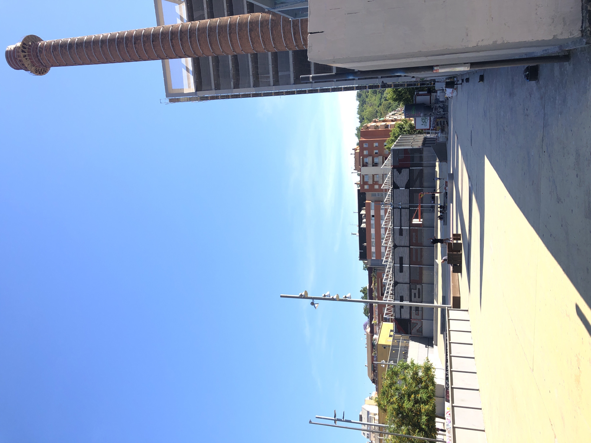 An outdoor, low-angle, wide shot captures a sunlit urban scene with a clear blue sky. In the foreground, a light-colored concrete plaza is bathed in sunlight, casting long shadows from a modern, angular structure on the right. The plaza extends towards the background, where it meets a large, open area with several benches and two figures. To the left of the plaza, a low, grey, rectangular barrier runs horizontally, lined with vertical posts and topped with what appears to be decorative fencing. Beyond this barrier, a large green tree stands tall, its branches full of leaves. Further in the background, a modern, multi-level building with a grey facade and open-air floors is under construction. Adjacent to this, a tall, cylindrical brick smokestack with horizontal banding dominates the right side of the image. Behind the construction and smokestack, a cluster of multi-story apartment buildings in various colors, including orange and yellow, are visible, nestled against a backdrop of green, tree-covered hills. Several lampposts with multiple lights are positioned throughout the scene, particularly on the left and in the mid-ground. The overall impression is one of an urban redevelopment or a new public space being established.