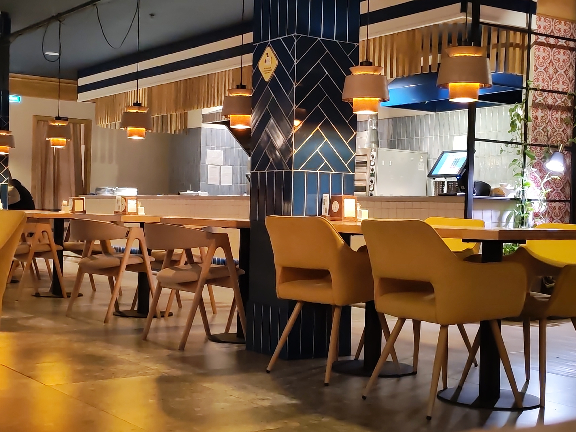 The image depicts a restaurant interior. Several wooden tables with chairs are arranged in a row. The chairs are of two types: wooden-framed chairs with upholstered seats and backs, and yellow upholstered chairs with wooden legs. Modern pendant lights hang from the ceiling, illuminating the space. In the background, a partial view of the kitchen is visible, with stainless steel equipment and tiled walls. The interior design incorporates a dark blue, herringbone-patterned pillar. The restaurant's ambiance is modern and well-lit. The image doesn't provide enough information to determine if the image is in Izhevsk, Russia.