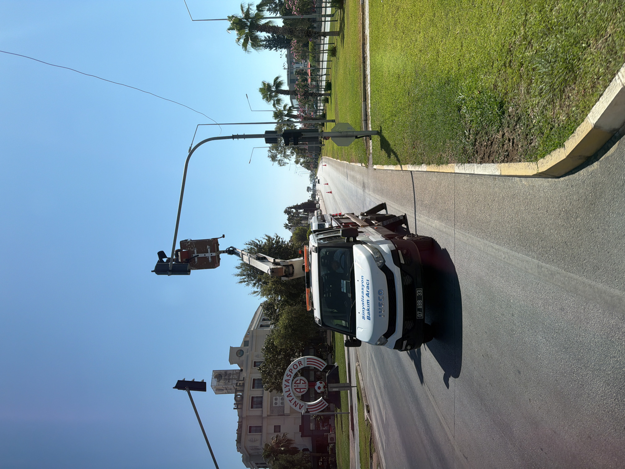 In the city of Konyaaltı, Turkey, a white Iveco maintenance vehicle, identified by the license plate "07 ABB 33", is parked on the right side of a road. The vehicle is equipped with a boom lift, currently extended and positioned near a traffic light. A person, wearing a dark shirt and possibly a hat, is visible in the driver's seat. The vehicle has "Sinyalizasyon Bakım Aracı" written on its side, which translates to "Signaling Maintenance Vehicle".
In the background, a large building with "HOTEL" signage is prominent. Adjacent to the hotel, a circular sign with "ANTALYASPOR" branding is displayed. The scene is illuminated by bright sunlight under a clear blue sky. The road has multiple lanes and appears to be a main thoroughfare. On the right side of the road, a well-maintained grassy area with palm trees and other vegetation lines the street, bordered by a sidewalk and a curb. Traffic cones are visible further down the road, suggesting potential ongoing work or traffic management. The overall environment indicates an urban setting on a sunny day.