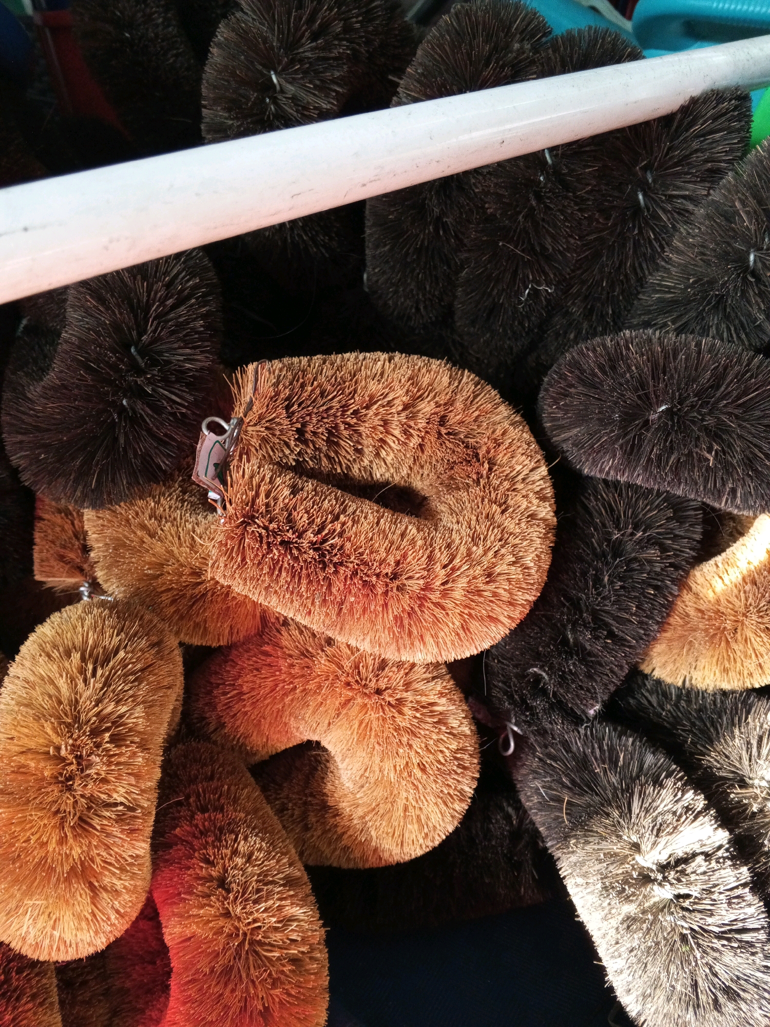 A pile of coconut fiber cleaning brushes is displayed. The brushes vary in color from light brown to dark brown and black. Some brushes are round and bushy, while others are elongated and curved. A white pipe runs horizontally across the top portion of the image, partially obscuring some of the brushes. The background is dark. There is no indication of the location being in Bintang Meriah, Indonesia.