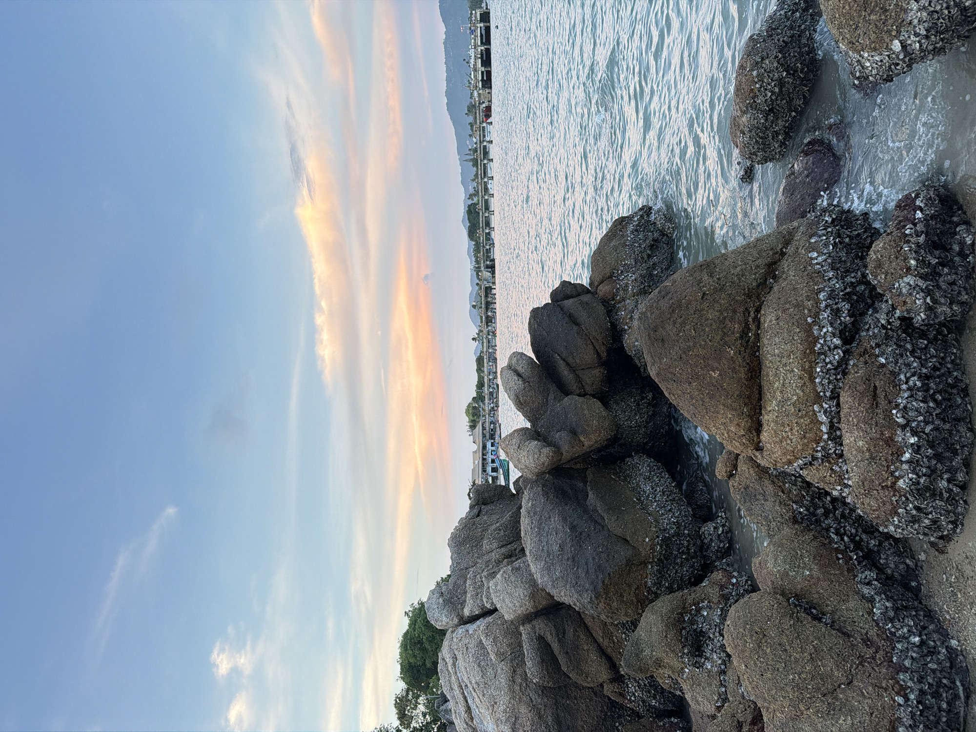 This is a horizontal, eye-level, medium shot of a rocky coastline at sunset in Ko Samui District, Thailand. The foreground is dominated by large, weathered rocks covered in barnacles, with the ocean lapping at their base. The rocks are dark gray and brown, with some patches of green moss. The water is a calm, turquoise color, reflecting the soft hues of the sky. In the background, a long pier stretches out into the water, lined with buildings and a few boats. Mountains are visible in the distance, silhouetted against the sunset. The sky is a gradient of soft blues, pinks, and oranges, with wispy clouds scattered across it. The overall mood is serene and tranquil. There are no people visible in the image.