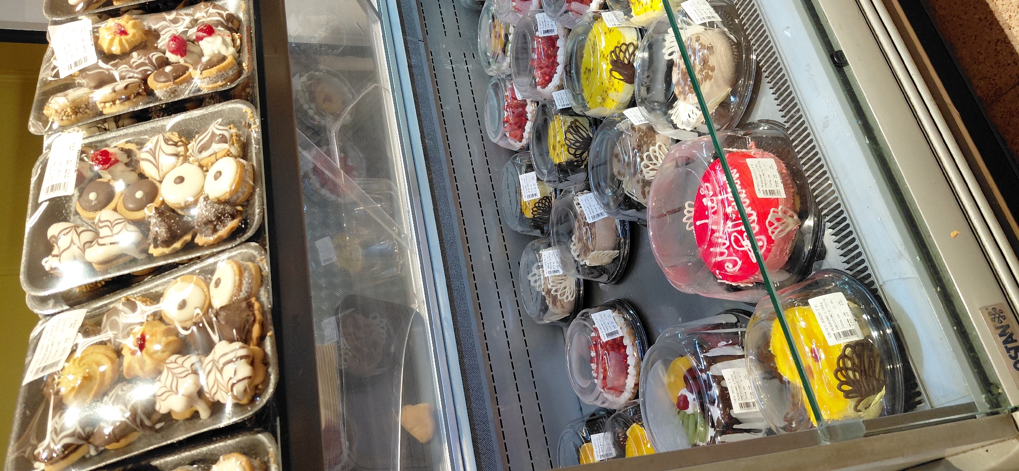 This image shows a display case in a bakery in Encarnación, Paraguay, filled with various types of cakes and pastries. The display case is well-lit, and the items are arranged in plastic containers with clear lids.
In the upper section of the display, there are trays of small cookies and pastries, some topped with chocolate drizzle, icing, or fruit. These appear to be packaged for sale in bulk.
The lower section features individual cakes. There are several round cakes with colorful frosting and decorations, including a red cake with white script lettering that says "Feliz Cumpleaños" (Happy Birthday). Other cakes have fruit toppings, chocolate decorations, or a marbled frosting. Some of these cakes have price and product information labels attached.
The environment suggests a retail setting, likely a supermarket or a dedicated bakery, with the products presented for customers. The overall impression is one of a well-stocked and inviting display of baked goods.