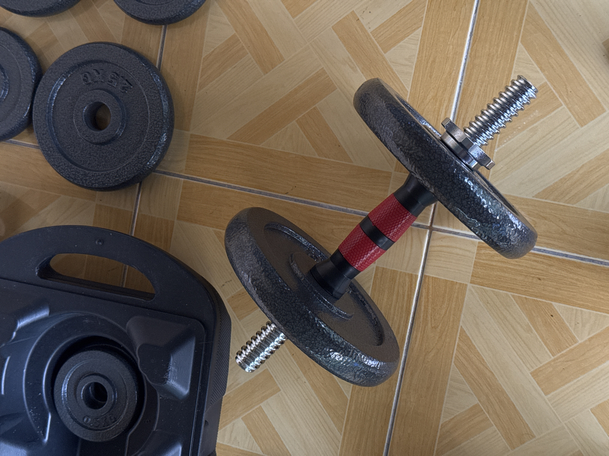 The image shows a dumbbell with two weight plates attached to it, lying on a tiled floor. The dumbbell has a black grip with red bands around the middle and a silver threaded end where the weight plates are attached. The weight plates are dark gray with a textured surface and a central hole. One of the plates has visible text that appears to read "2.5 KG". To the left of the dumbbell is a black plastic case, likely for storing dumbbells, with a weight plate nestled inside. The case has a handle and a contoured interior. The floor is tiled with a pattern of light and dark beige rectangular tiles arranged in a parquet style. The lighting suggests an indoor setting, possibly a home gym or workout area. No people are visible in the image. The scene is still and focused on the weightlifting equipment.