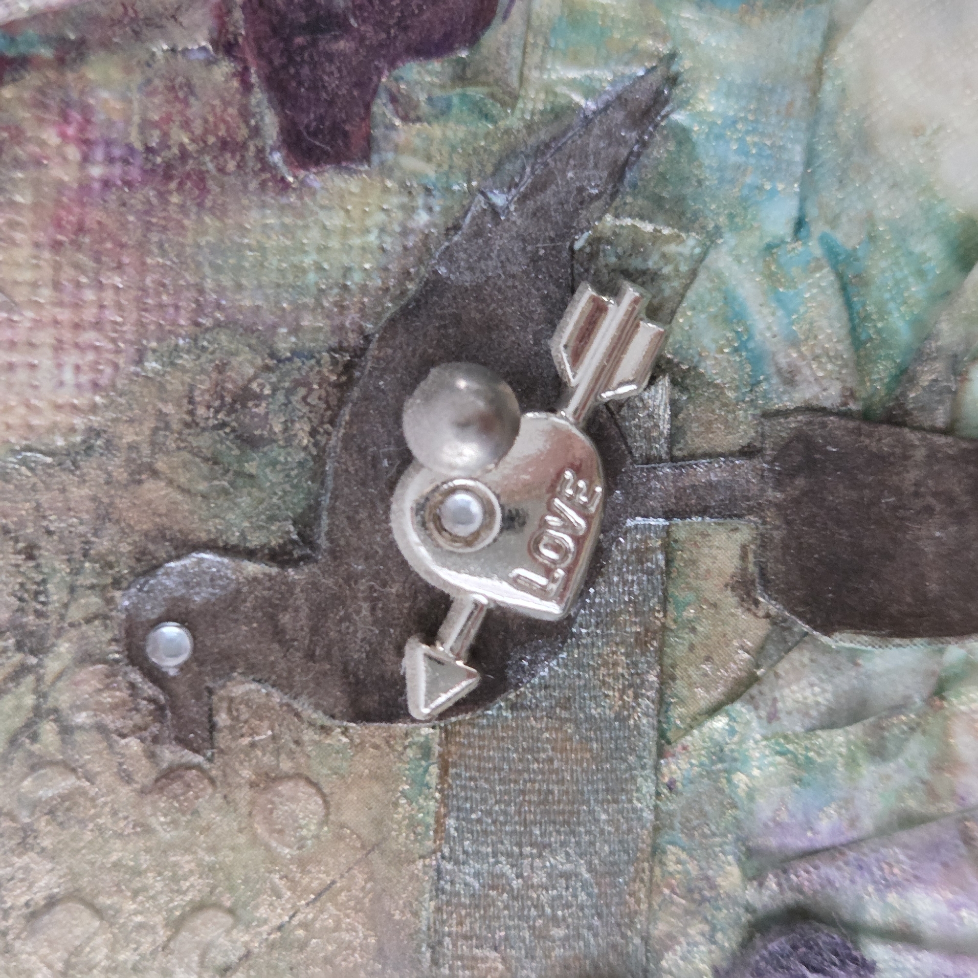 A close-up photograph depicts a detail from a mixed-media art piece, focusing on a metallic charm against a textured, colorful background.
The central object is a silver-toned metallic charm, shaped like a heart pierced by an arrow. The word "LOVE" is clearly inscribed in raised capital letters on the heart's surface. The charm is secured by a small central rivet and a larger, domed silver bead above it. This heart-and-arrow charm is affixed to a dark, possibly metallic or painted, bird-shaped cutout, which resembles a dove or swallow. The bird shape is itself attached to the background with at least two visible silver rivets, one near its head and another partially visible beneath the charm.
The background consists of a richly textured surface, likely fabric or canvas, covered with layers of paint, ink, and possibly glitter or iridescent mediums. A vibrant array of colors is present, including shades of pink, purple, teal, light blue, green, and earthy browns, with a noticeable golden or silvery shimmer dispersed throughout. The texture is varied, showing visible brushstrokes, fabric weaves, and small, raised elements or bubbles. The intricate layering and varied textures suggest a handcrafted or artistic creation. No specific activity or event is taking place beyond the static display of the artwork. The dominant theme, conveyed by the heart, arrow, and inscribed "LOVE," is one of affection or romance.
