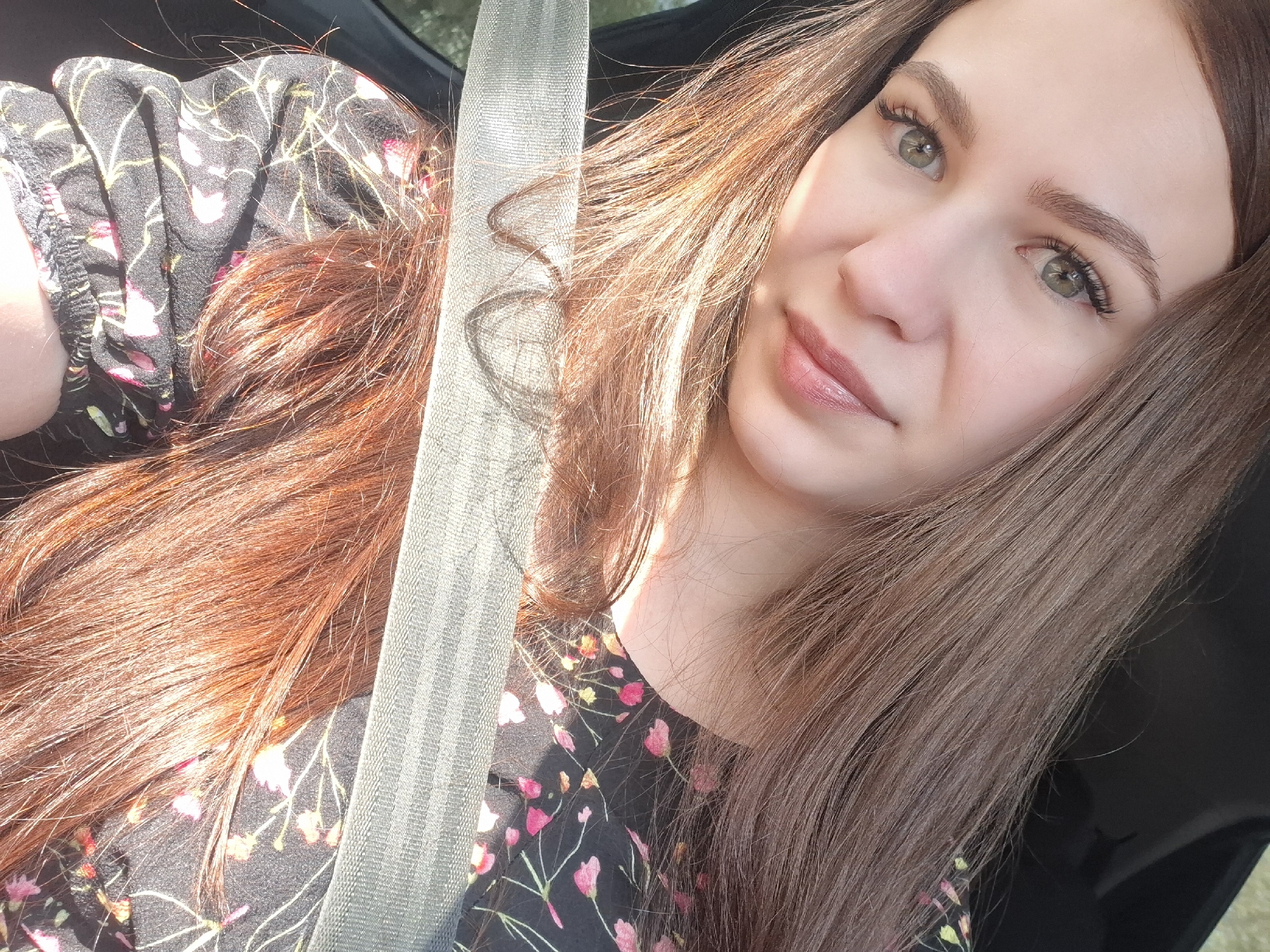 A young woman is taking a selfie indoors, possibly in a car, as she is wearing a seatbelt. She has light skin, long light brown hair, and green eyes. She is wearing a dark-colored floral print dress. The location is not identifiable as Chaikovsky, Russia, without additional information.