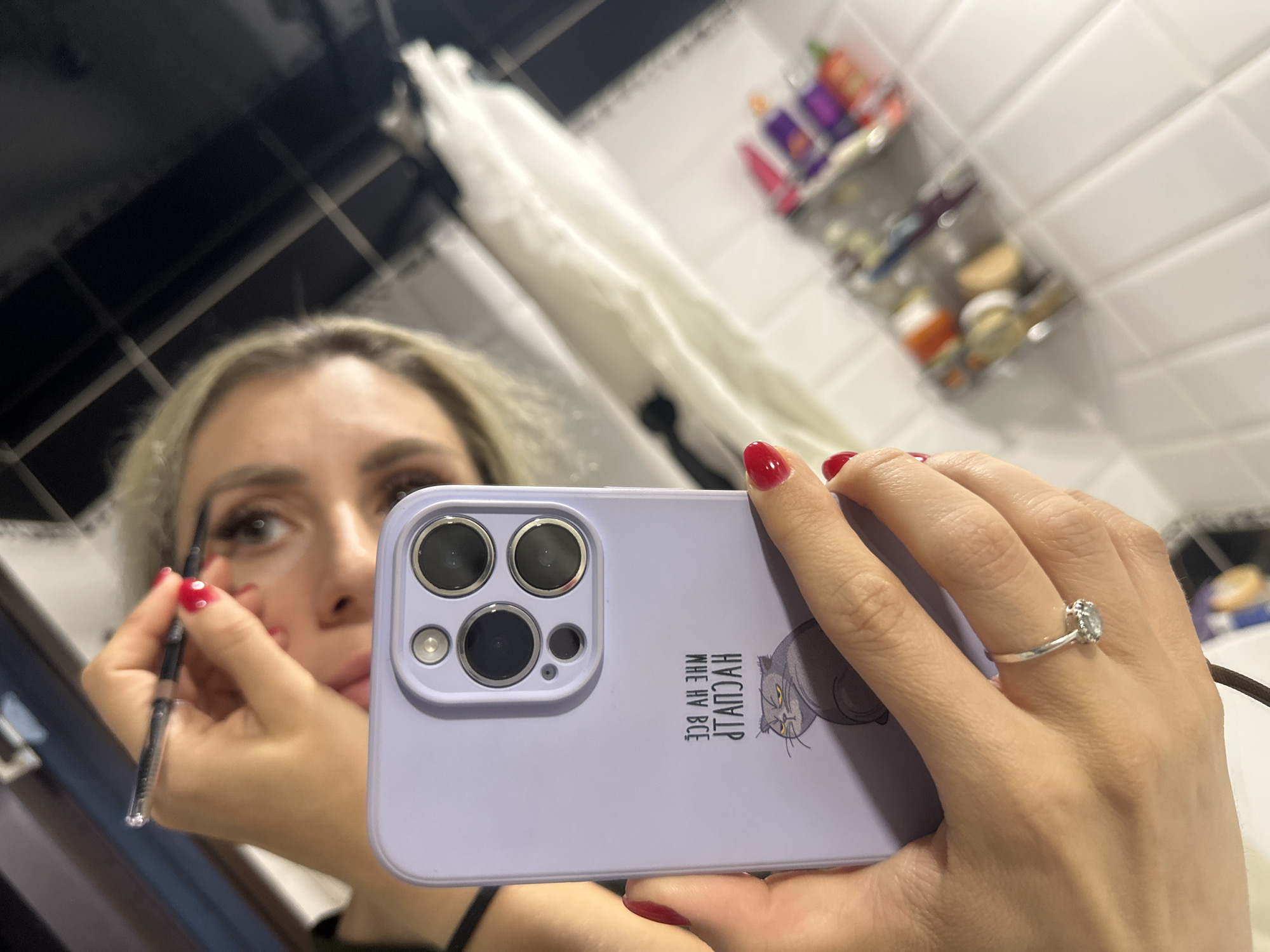 A woman with blonde hair is applying makeup in a bathroom, as seen in a reflection. She is holding a makeup pencil to her left eyebrow with her right hand. Her hand is adorned with a silver ring with a diamond. Her fingernails are painted red. She is holding a lavender iPhone in front of her, obscuring part of her face. The phone case has a picture of a cat and text in Russian that appears to read "Я Кот" which translates to "I am a cat." In the background, there are shelves with various toiletries and what looks like a shower curtain. The lighting suggests it is daytime. The setting appears to be a residential bathroom in Novoaltaysk, Russia.