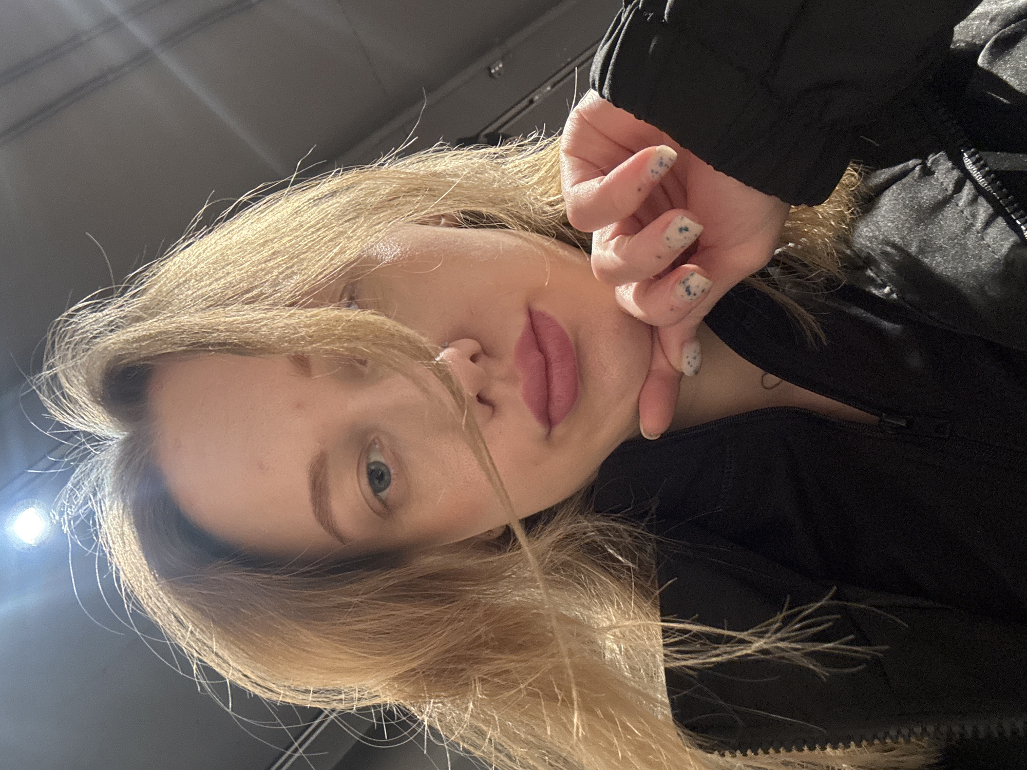 A close-up, low-angle selfie of a fair-skinned young woman with blonde hair. She is wearing a dark, possibly black, jacket. Her hand with nails painted in a light color with a blue speckled design is touching her face. The environment is unclear, with only a glimpse of a possibly gray ceiling and a light fixture visible in the background. The image provides no information to identify the location as Rostov-on-Don or any other specific place.