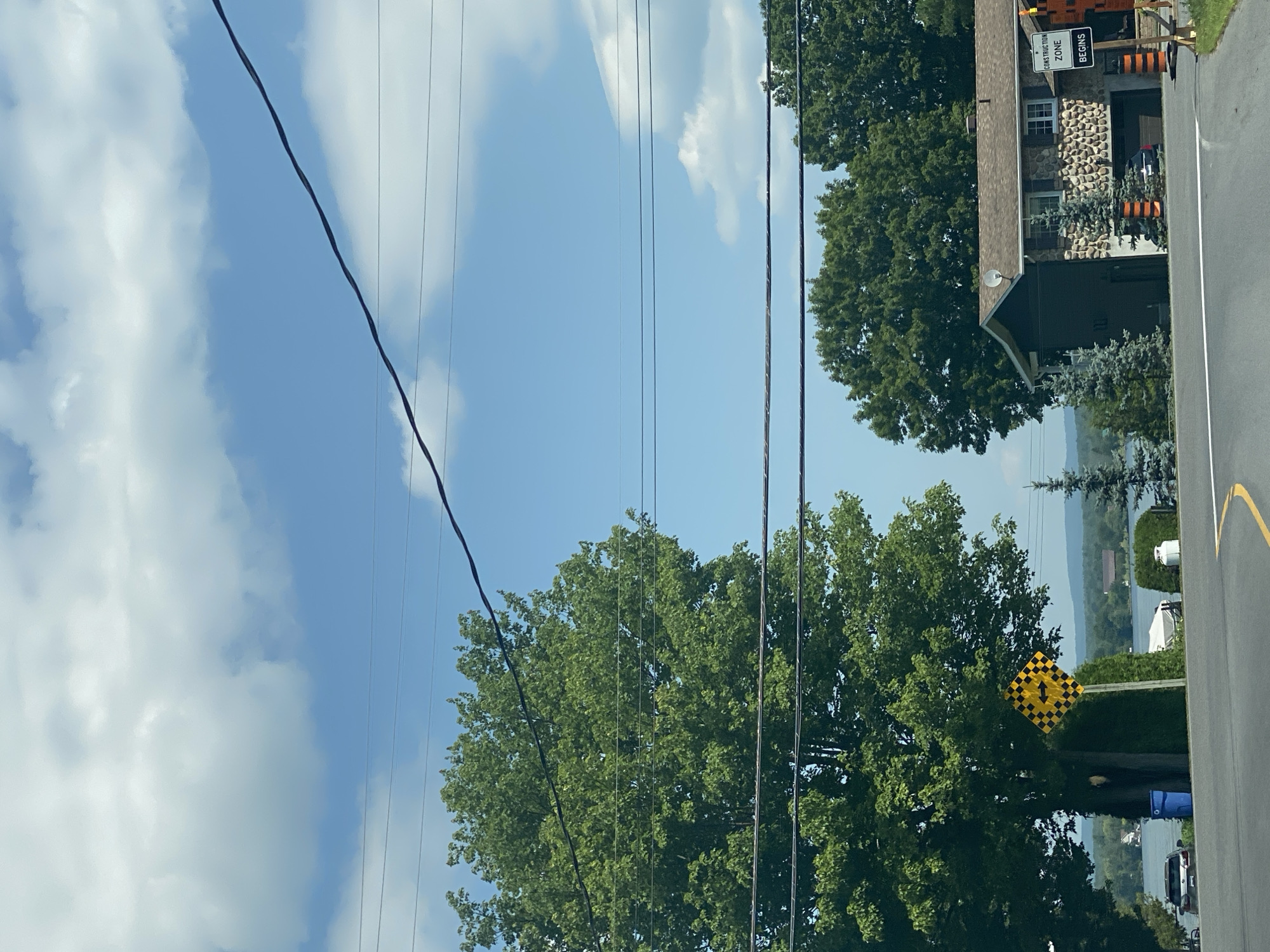 This is a ground-level, eye-level, static, medium shot photograph taken in East Hawkesbury, Canada, on a sunny day with scattered clouds. The image captures a residential street scene bordering a body of water. In the foreground, a paved road curves to the left. A large deciduous tree with lush green foliage dominates the left side of the frame, partially obscuring a blue trash bin and a parked silver sedan. To the right of the tree, a diamond-shaped traffic sign with a black and yellow checkered pattern and a black arrow pointing left is mounted on a pole.
Further into the scene, a two-story house with a stone and dark siding exterior is visible. The house has a dark-colored garage door and several windows. In front of the house, small evergreen trees with orange striped bases are planted. A white propane tank sits on the right side of the yard. A boat is moored near the water's edge.
Beyond the house and trees, a wide expanse of water is visible, with a distant shoreline featuring more trees and buildings. Power lines stretch across the sky, intersecting the scene. A sign indicating "CONSTRUCTION ZONE BEGINS" with orange traffic cones is present near the right edge of the frame, suggesting ongoing road work. The overall impression is one of a peaceful, semi-rural residential area.