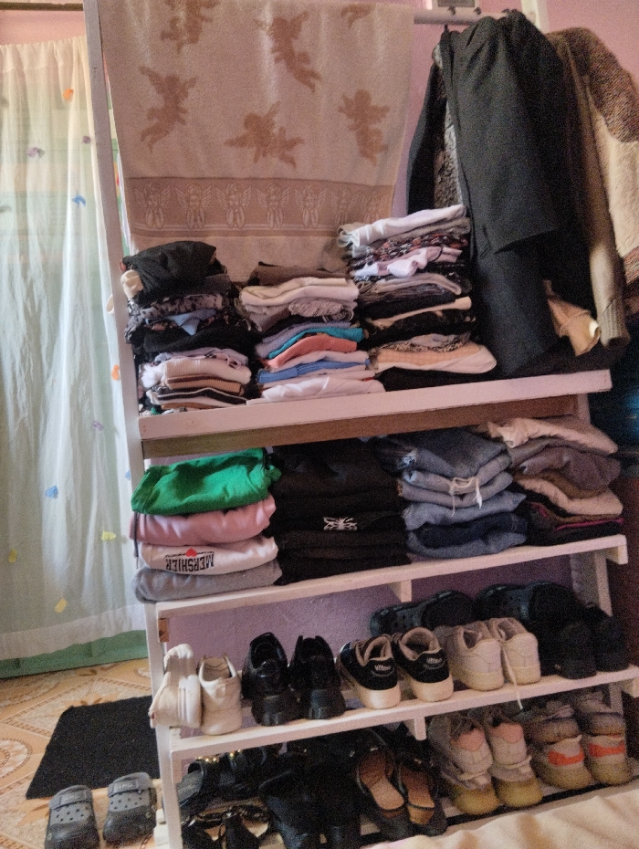 An indoor scene in Witeithie ward, Kenya, depicts a three-tiered white wooden shelving unit organized for clothing and footwear storage.
On the topmost rail, a light-colored blanket or towel featuring repeating cherub motifs in brown hangs draped. To its right, dark garments are suspended from hangers, with another lighter textured item partially visible.
The top shelf contains three distinct stacks of folded apparel: a mixed dark and light stack on the left, a predominantly light-colored stack in the center including white, blue, and pink items, and a mixed dark and light stack on the right.
The middle shelf also holds three stacks: a green garment atop a pink and a grey item (the grey item bearing "MERSHIER" text) on the left, dark-colored items in the center (one with a spider-like emblem), and folded blue denim garments atop other items on the right.
The bottom shelf is dedicated to footwear, displaying from left to right: two pairs of white sneakers, two pairs of black platform shoes, two pairs of dark-colored low-top sneakers, and two pairs of dark clogs.
On the tiled floor in front of the unit, two pairs of dark grey Crocs-style clogs are present. Further pairs of shoes, including black flats, heels, and multi-colored sneakers, are visible on the floor beneath and around the base of the shelving unit.
The background features a translucent white curtain adorned with small, colorful shapes on the left. The wall behind the unit is a light, possibly lavender hue. The floor is tiled with a light brown pattern, and a small black mat rests on the tiles.