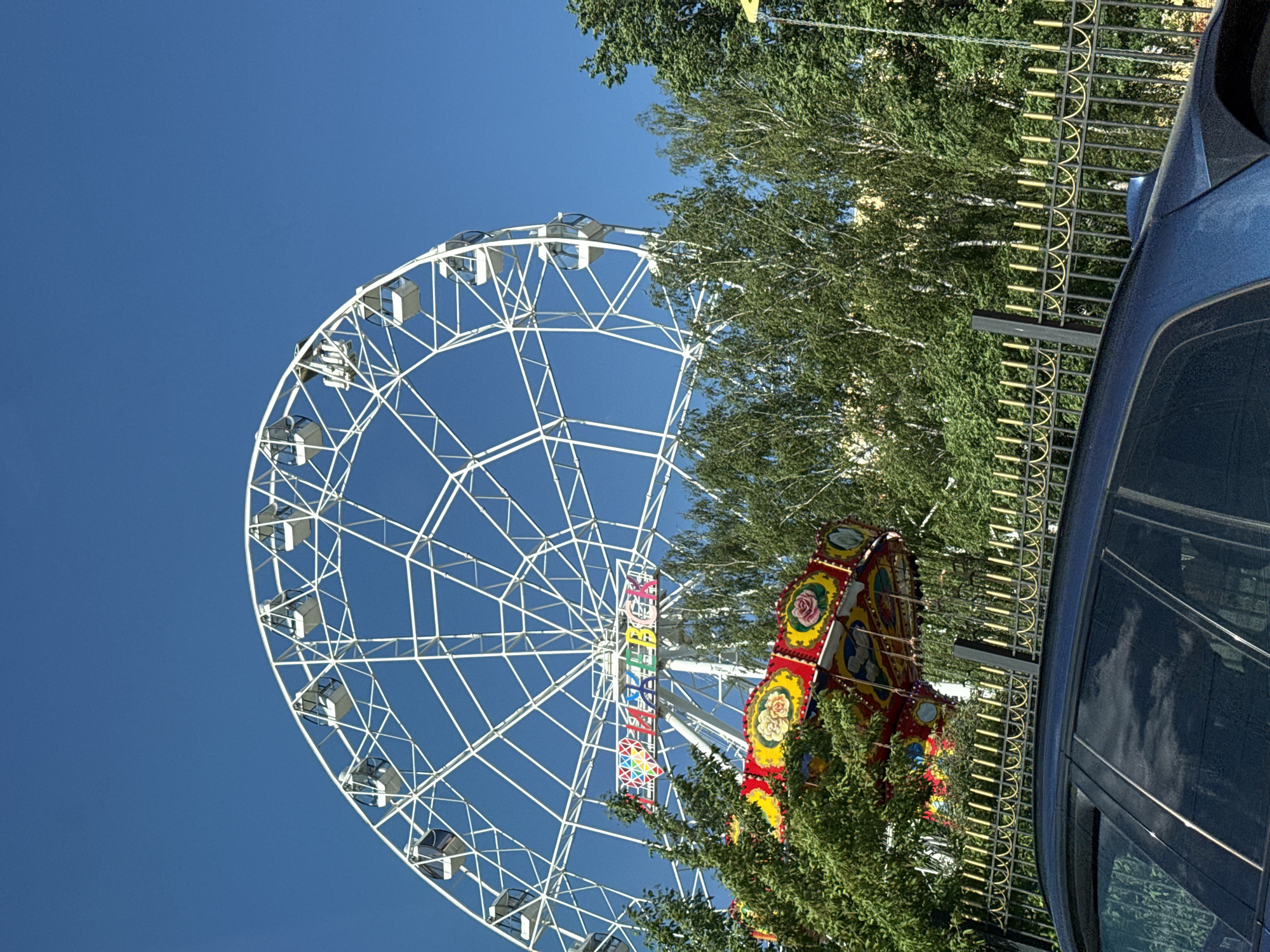 A low-angle, daytime shot showcases a Ferris wheel dominating the left side of the frame against a clear blue sky. The Ferris wheel is white and features numerous enclosed passenger cars. Adjacent to the Ferris wheel is a brightly colored carousel-style ride. The rides are positioned in a park area with trees and a metal fence. A portion of a dark-colored vehicle is visible on the right side of the image, reflecting the sky. The photo depicts the amusement rides in Izhevsk, Russia.