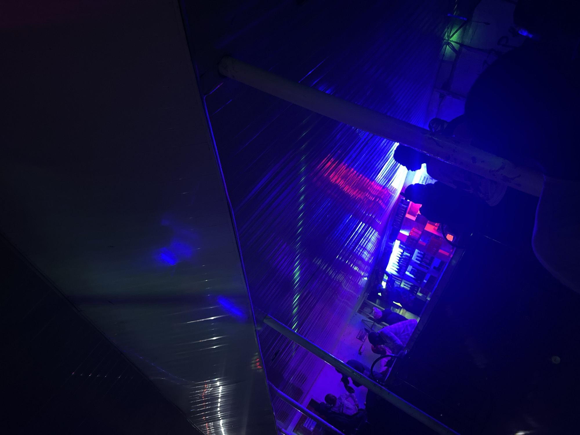 The image depicts an indoor scene at a bar or lounge in Wumat, Nigeria, bathed in vibrant blue and purple lighting, with streaks of red and green light reflecting off the corrugated ceiling. Silhouetted figures are visible, some standing near a bar that appears stocked with bottles. The overall atmosphere suggests a lively, nighttime setting, possibly a nightclub or entertainment venue.