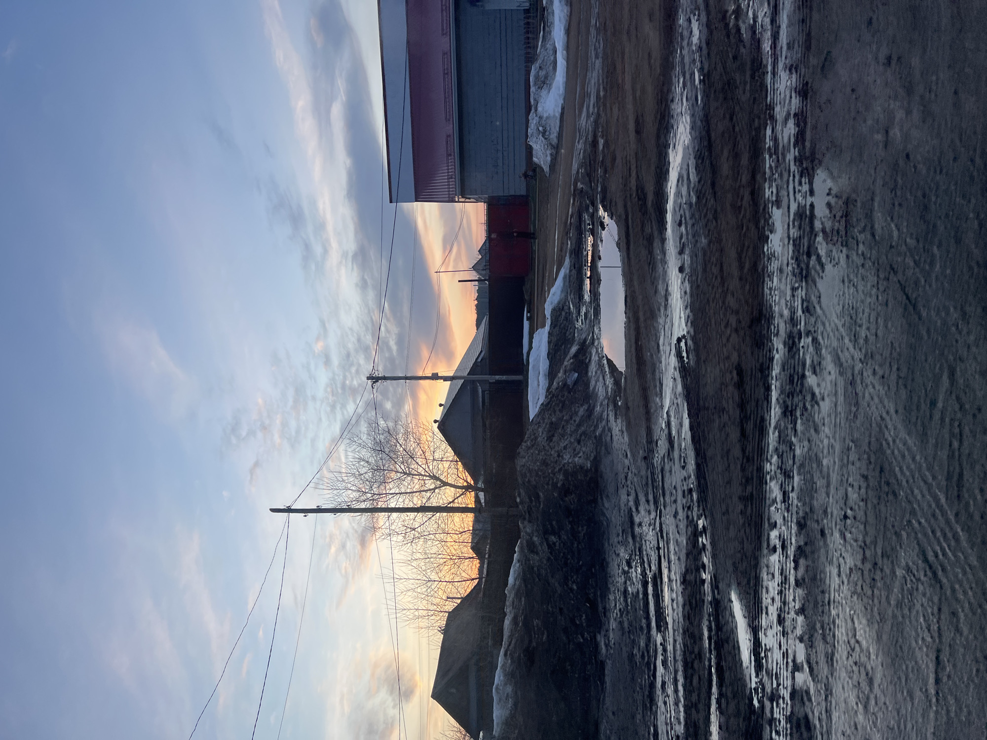 The image depicts an indistinct location in Chaikovsky, Russia. It appears to be a street or alleyway, possibly during or after a thaw, as evidenced by the mud, ice, and melting snow. Several buildings line the street, with one appearing to have a red and white roof. Power lines run across the sky, which shows a sunset or sunrise. A bare tree stands in the middle ground. The ground is uneven, covered in a mix of mud, ice, and tracks, likely from vehicles. The overall scene suggests a rural or semi-urban setting during a transitional season.