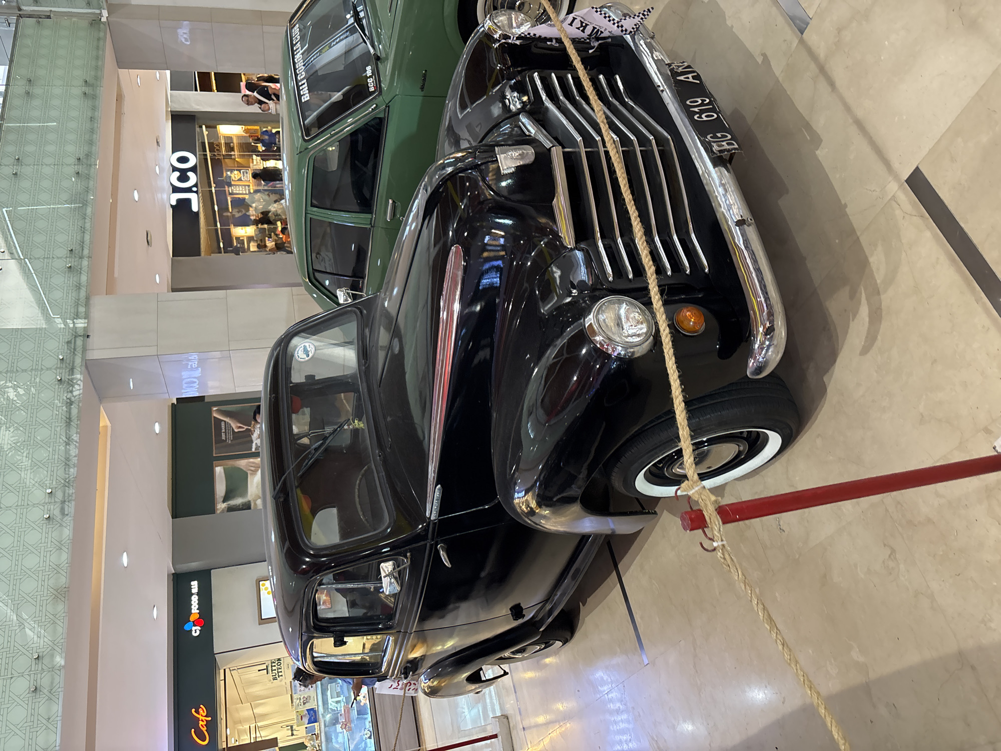 The image depicts two classic cars, one black and one green, on display in an indoor environment. The black car is in the foreground, while the green car is in the background. Both cars are cordoned off by a red pole and rope barrier. In the background, there are shops and eateries visible, including a "J.CO" store and "CJ FOOD LLE". The number plate "BG 619 AR" is visible on the front of the black car. Additionally, the green car features a "BALI COROLLA CLUB BCC 106 MKI" license plate. The scene appears to be within a shopping center or similar commercial complex in Kuta, Indonesia.
