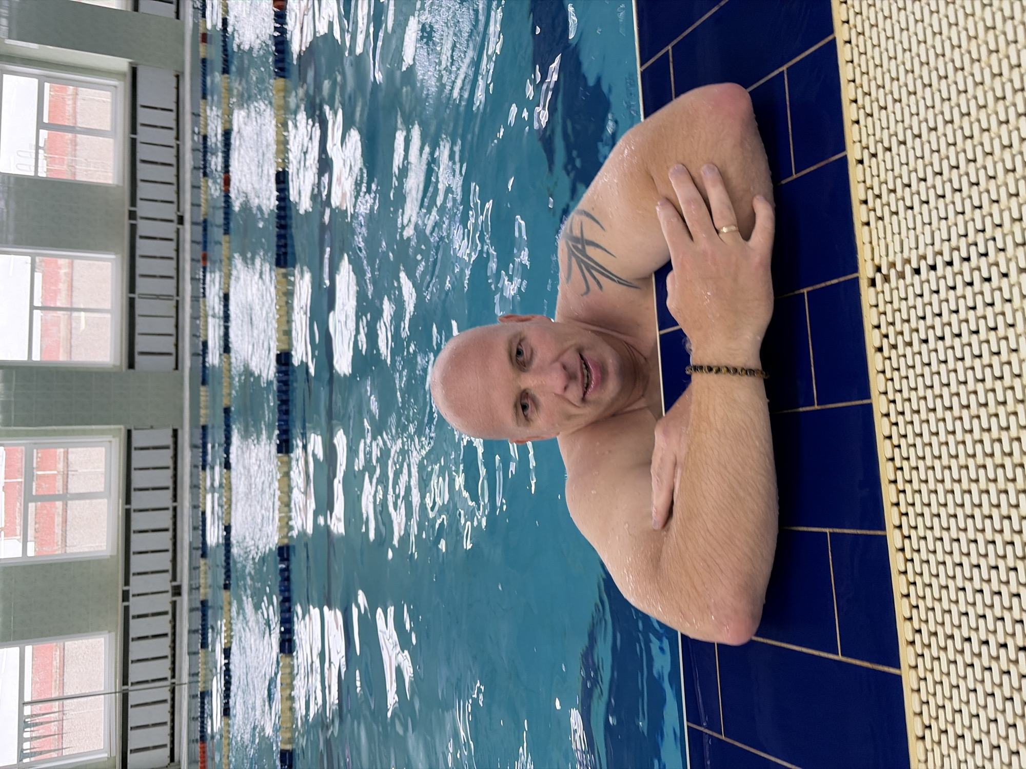 A man is in a swimming pool, leaning against the edge. He is bald and has a tattoo on his left arm. He is smiling and looking at the camera. The pool has lane markers and there are windows in the background. The man is wearing a ring on his left hand and a beaded bracelet on his right wrist. The water in the pool is blue and appears to be clean. The man's skin is wet, indicating he has been in the water. The scene is likely indoors, given the presence of windows and the tiled wall. The overall mood is casual and relaxed.