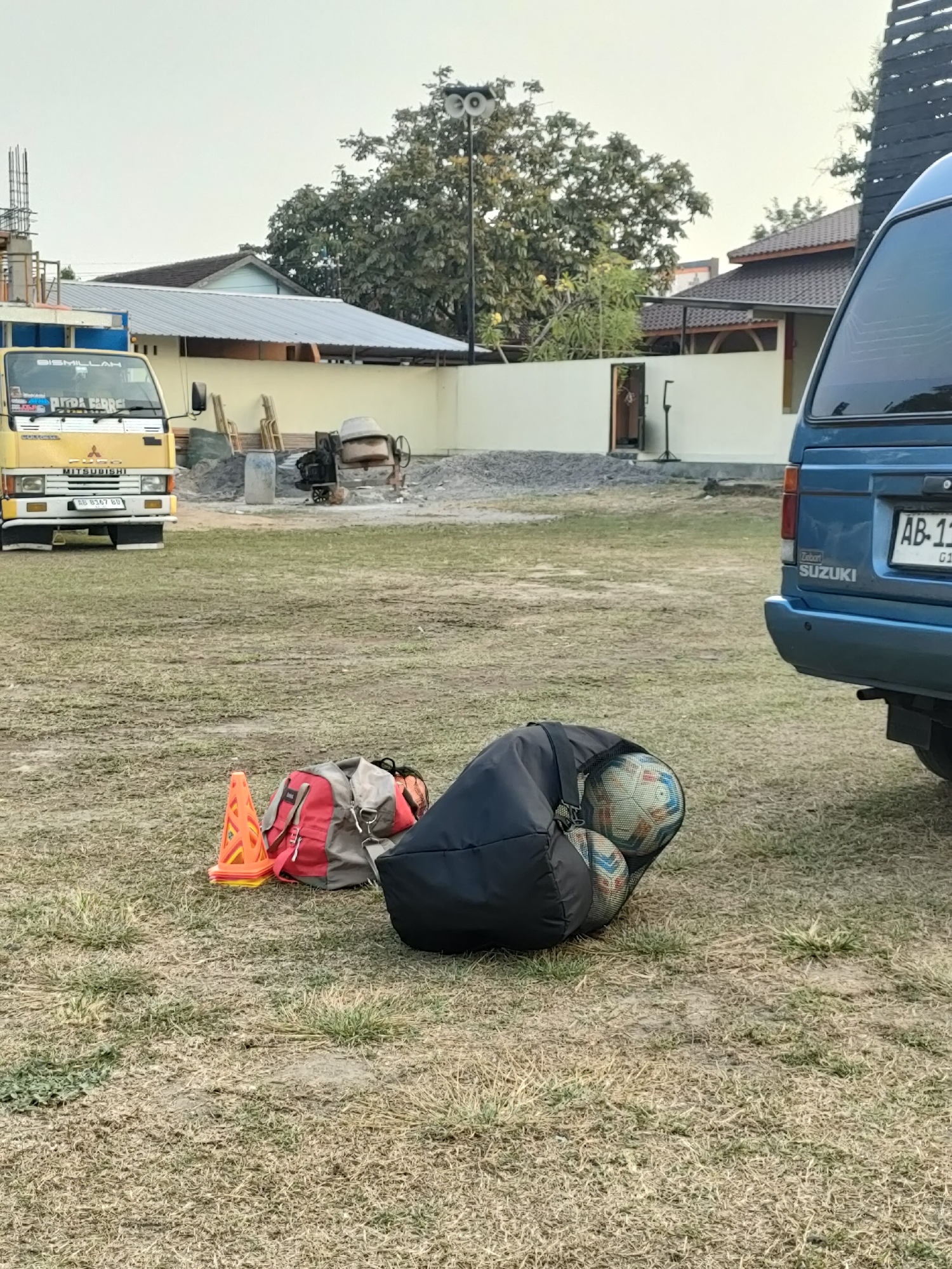 In Banguntapan, Indonesia, a scene unfolds in an outdoor lot, possibly a construction or event preparation area. In the foreground, a black duffel bag rests on dry, patchy grass, containing two soccer balls, one of which is partially visible and displays a pattern of multicolored panels. Adjacent to the bag, a stack of orange traffic cones, one with a yellow top, stands upright. Behind the cones, a red and gray backpack is placed on the ground, with straps visible.
Further into the background, a yellow Mitsubishi truck is parked, its front end facing the viewer. The truck has a visible license plate that reads "AB 6167 BD". To the right of the truck, a concrete mixer sits on a pile of gravel. A white wall stretches across the midground, with a light blue building visible beyond it. The sky above is overcast and pale. To the far right, the rear section of a blue Suzuki van is parked, its license plate showing "AB-11". The overall environment suggests an area undergoing work or preparing for an activity, with elements of construction and sports equipment coexisting.