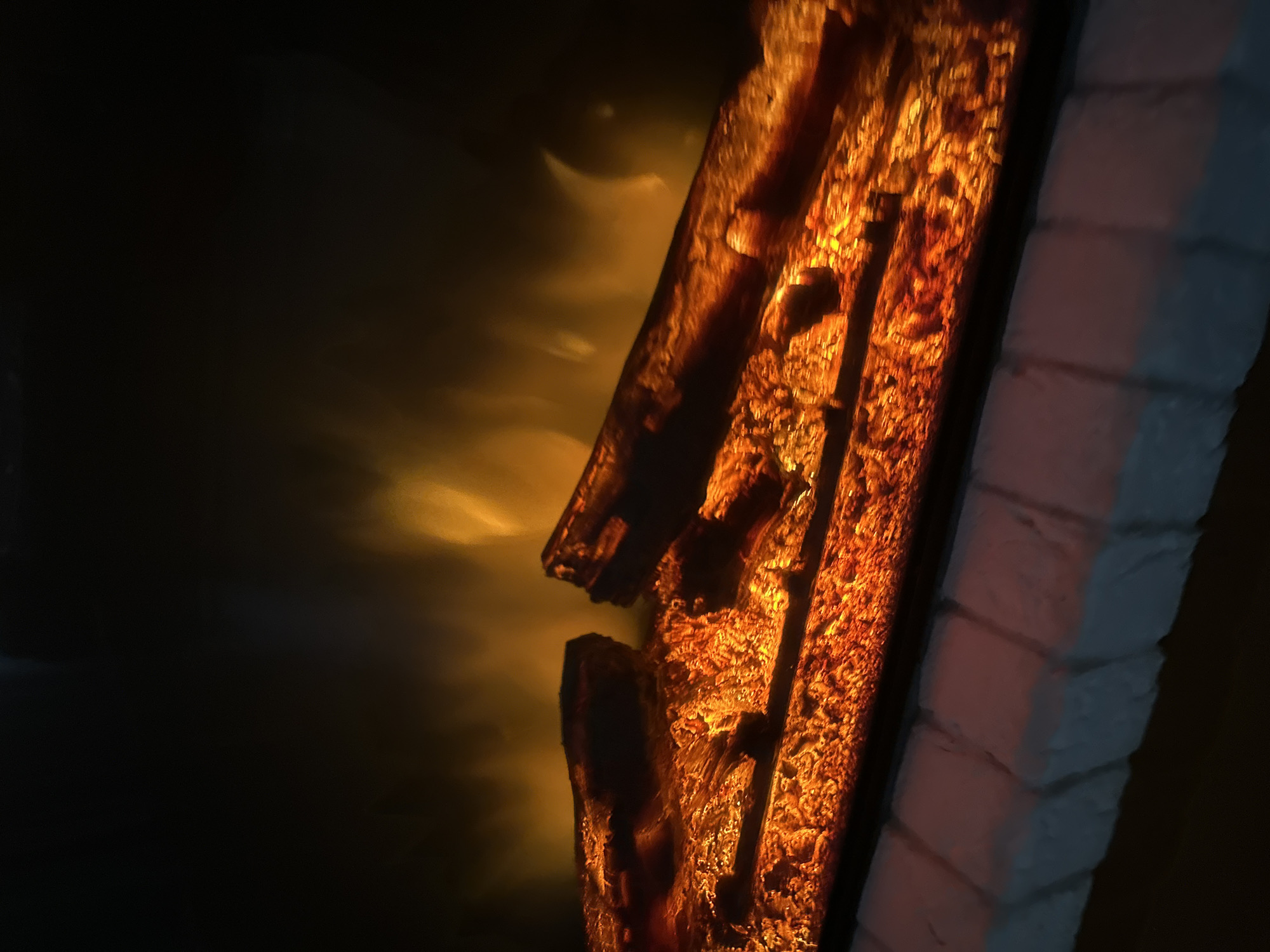 The media file is a close-up, angled shot of an artificial fireplace with flickering orange and yellow flames. The flames are located on the right side of the image, illuminating a textured, dark brown object that resembles burning logs. The texture of the logs is rough and irregular, with visible cracks and variations in color, suggesting embers and heat. To the right of the logs, there is a section of white brickwork, also illuminated by the firelight, which appears to be part of the fireplace surround. The bricks are laid in a horizontal pattern.
To the left of the fireplace logs, the scene transitions to darkness, with soft, ethereal wisps of orange and yellow light appearing in the background. These luminous effects suggest smoke or reflections from the fire. The overall mood is warm and inviting, evoking the feeling of a cozy fire. The lighting is dramatic, with strong contrasts between the illuminated elements and the dark background. There is no visible text in the image. The time of day is indeterminate due to the artificial lighting and dark surroundings, but the scene suggests an indoor setting, likely a living room or similar domestic space. The image does not contain people.