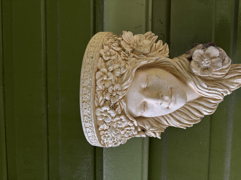 A cream-colored, weathered wall planter, sculpted as the head and upper torso of a female figure, is mounted centrally against a dark olive-green wooden plank wall. The figure faces forward with eyes closed, a subtle smile, and long, wavy hair adorned with a large flower on its right side. A detailed floral wreath forms a crown around her head, topped by an ornate decorative band, collectively serving as the planter's rim. Traces of dirt, discoloration, and fine cobwebs are visible on the sculpture, particularly within the crevices of the hair and flowers, and on the decorative band. The background consists of horizontally aligned, painted wooden planks exhibiting some wear and faint cobweb presence on the left side. The scene is located in the city of Stiperstones, United Kingdom.