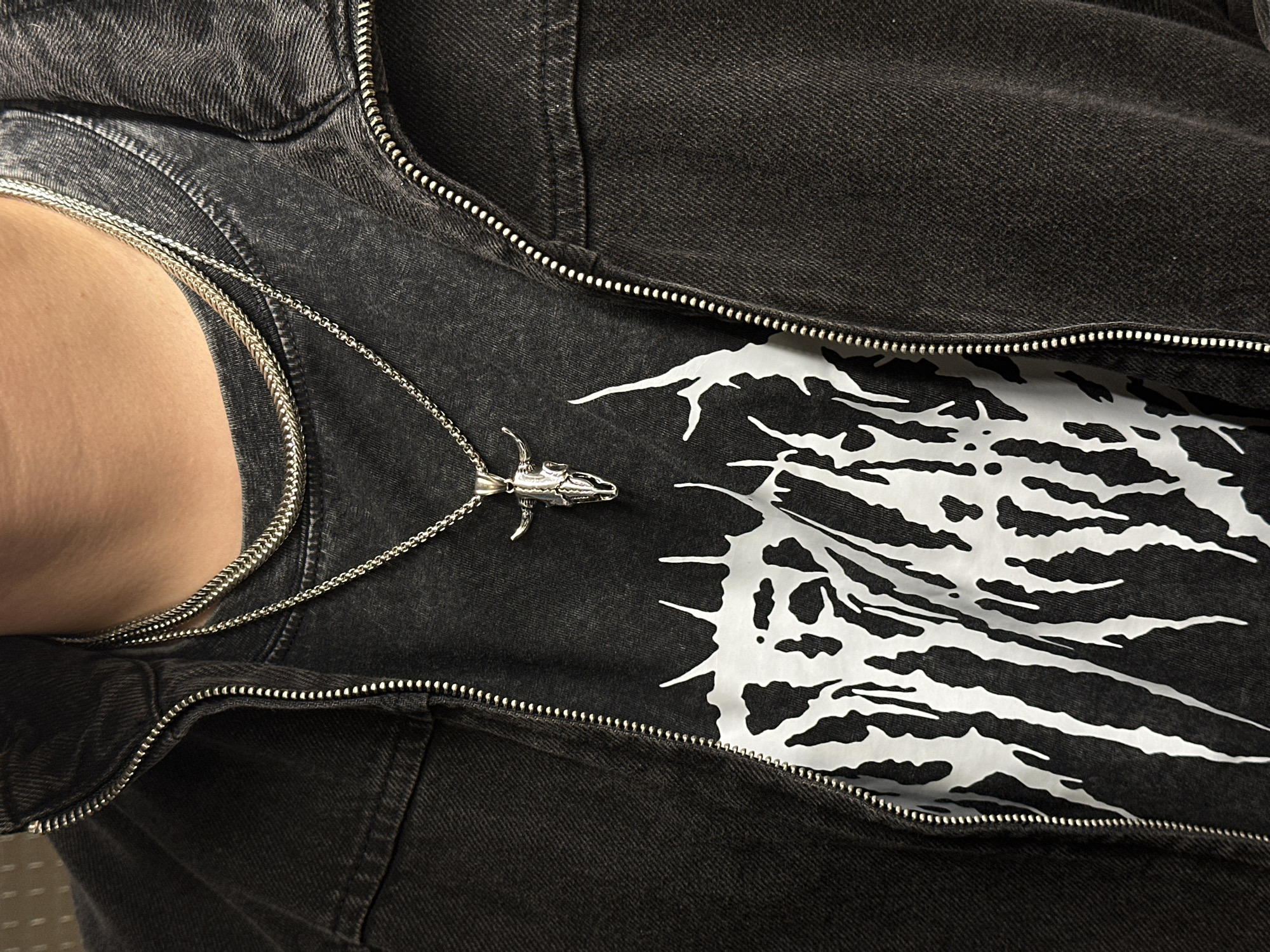 The image depicts a close-up view of a person's upper torso, adorned with clothing and jewelry. The individual is wearing a dark, possibly denim jacket, which is partially zipped up. Beneath the jacket, a darker t-shirt with a white graphic design is visible. A silver-colored necklace with a unique pendant, possibly depicting an animal skull, rests on the t-shirt. The person's skin is visible at the neck.
The photograph provides no information about the City of Zagreb, Croatia. Therefore, I can not fulfill the request to provide a location in that city.