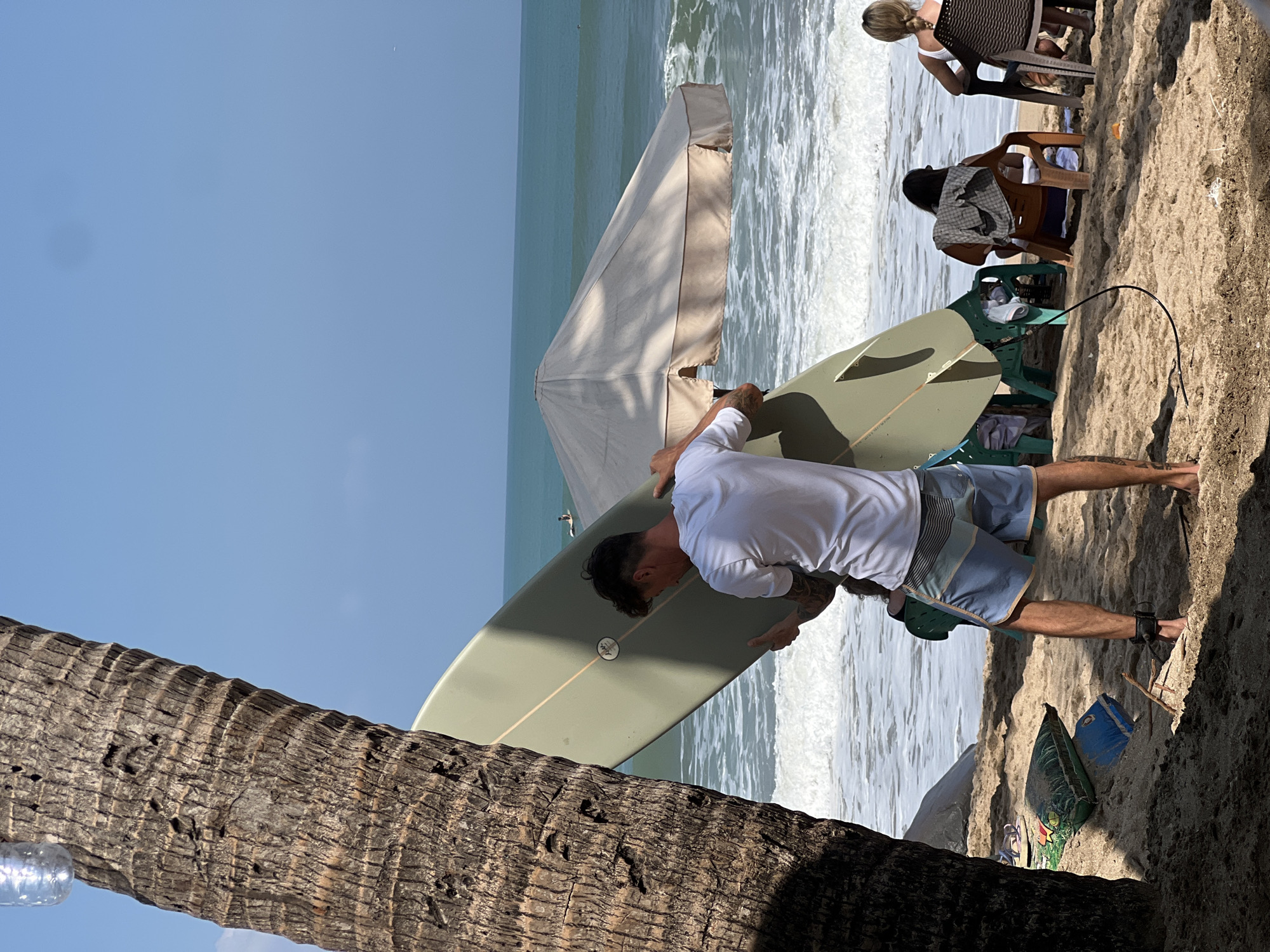 This image depicts a sunny beach scene in Kuta, Indonesia. A man, wearing a white t-shirt and blue patterned shorts, is holding a light green surfboard upright. He is standing near a palm tree on the sandy beach, with the ocean in the background. Waves are breaking on the shore, and the water appears turquoise. A large beige beach umbrella is set up on the sand, and several people are seated in chairs under it. In the distance, two surfers are visible in the water. There are also some beach bags and a piece of driftwood on the sand. The sky is clear and blue.