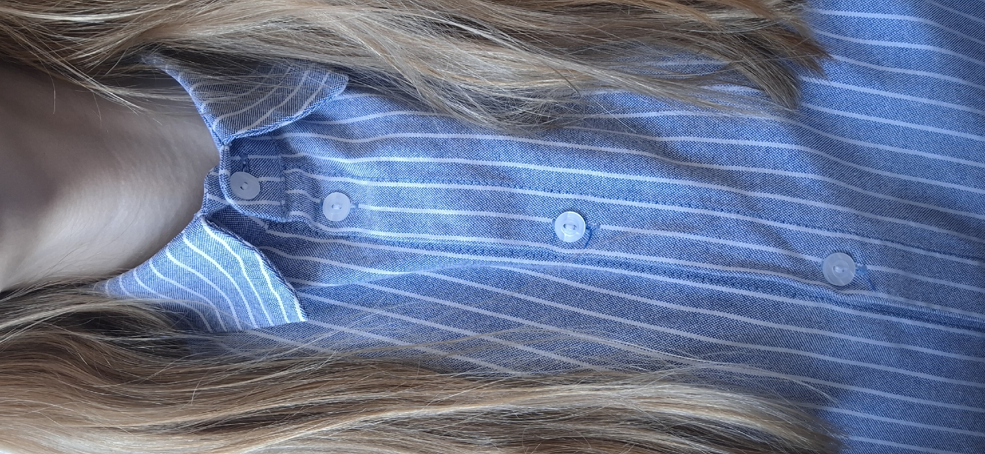 A close-up vertical image displays the upper chest and neck of an individual, with the scene located in Chaikovsky, Russia. Long, light-blonde hair frames the view, falling over both shoulders and partially obscuring a light blue button-down shirt. The shirt features thin, vertical white pinstripes. Three light-colored, possibly white or translucent, four-hole buttons are visible on the shirt's placket, with the topmost button beneath the open collar remaining unfastened. The skin of the upper neck and clavicle area is exposed above the shirt collar. No other individuals, actions, interactions, or environmental elements beyond the subject's immediate clothing, hair, and skin are discernible within the frame.
