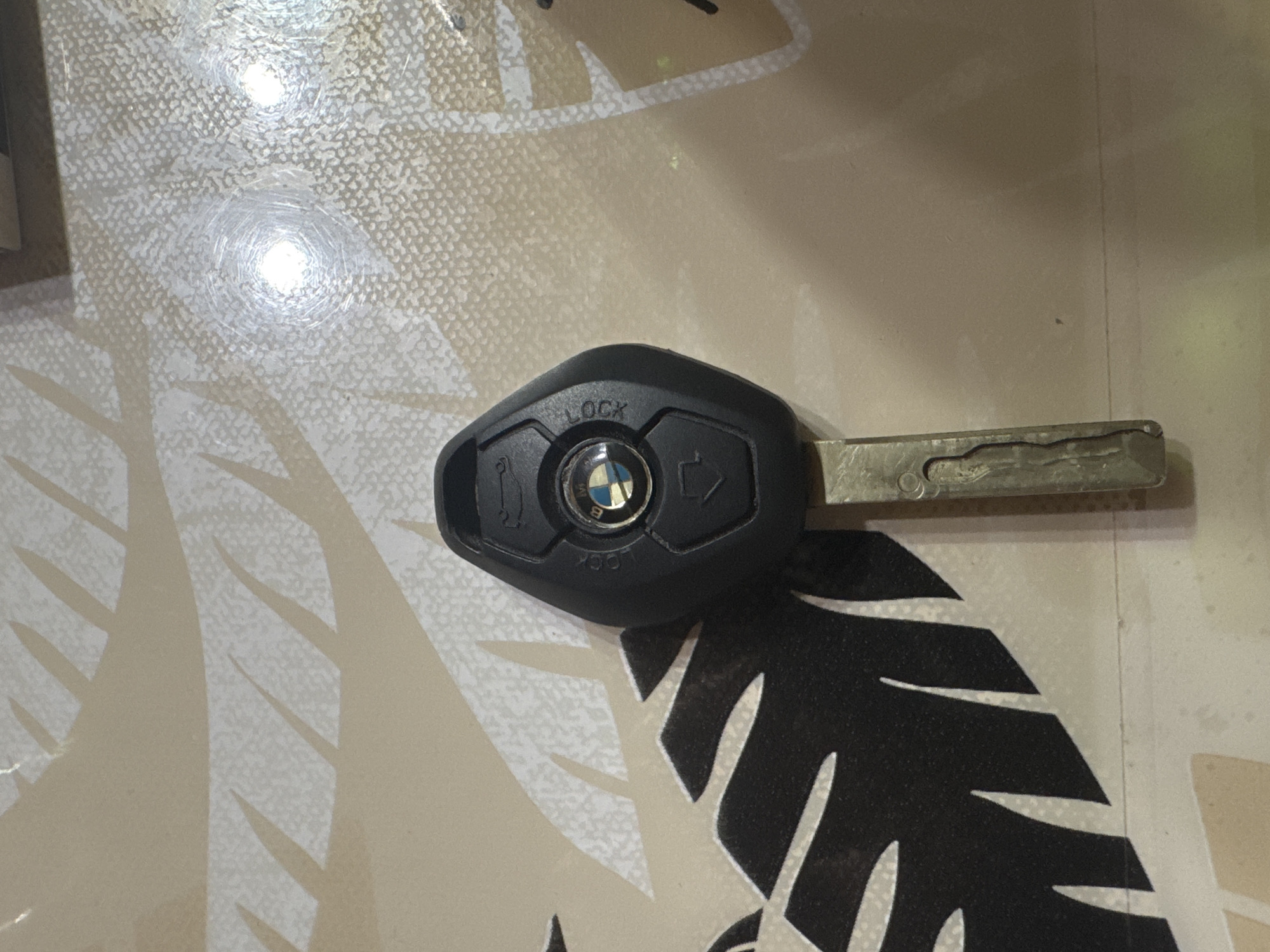 The image captures a single black BMW car key lying on a textured, patterned surface. The key fob is black, with a central circular area featuring the iconic blue and white BMW logo. Above the logo, the word "LOCK" is embossed. To the left of the logo, an open padlock symbol indicates the unlock function, and to the right, an icon resembling a car's trunk is visible. A metallic key blade extends from the right side of the fob. The key blade shows considerable signs of wear, including scratches and some tarnishing, suggesting frequent use.
The surface on which the key rests appears to be a countertop or table. It features a light beige or off-white base with a fine, mottled texture. Overlaid on this background are larger, abstract patterns resembling dark brown or black leaves or branches. Two distinct, bright circular reflections are visible on the surface, indicating strong light sources overhead or nearby, creating glare.
No individuals or other objects are present in the frame. The scene is static, with no discernible activity taking place. The key appears to be a functional car key, possibly for an older model BMW given the wear on the blade. The setting is indoors in Sarnevo, Bulgaria.