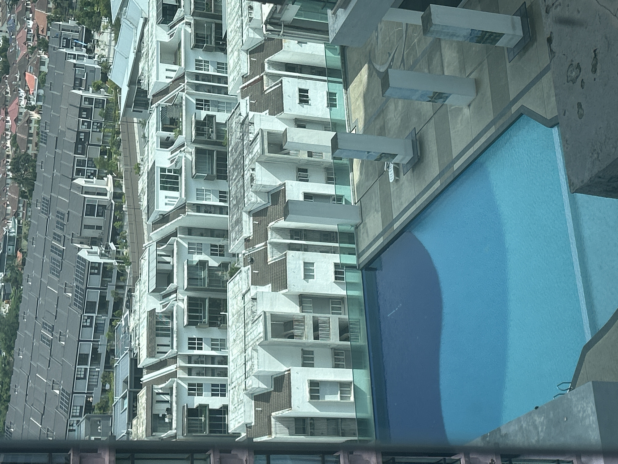 An aerial shot shows a dense residential area in Kuala Lumpur, Malaysia, featuring a mix of terraced houses with gray roofs on the left and modern white condominium buildings with balconies on the right. The terraced houses exhibit uniform architecture, while the condominium buildings display staggered heights and various design elements. A bright blue swimming pool occupies a corner of the frame, adjacent to the condominium complex. The photograph appears to have been taken from an elevated position, possibly from a higher floor of another building.