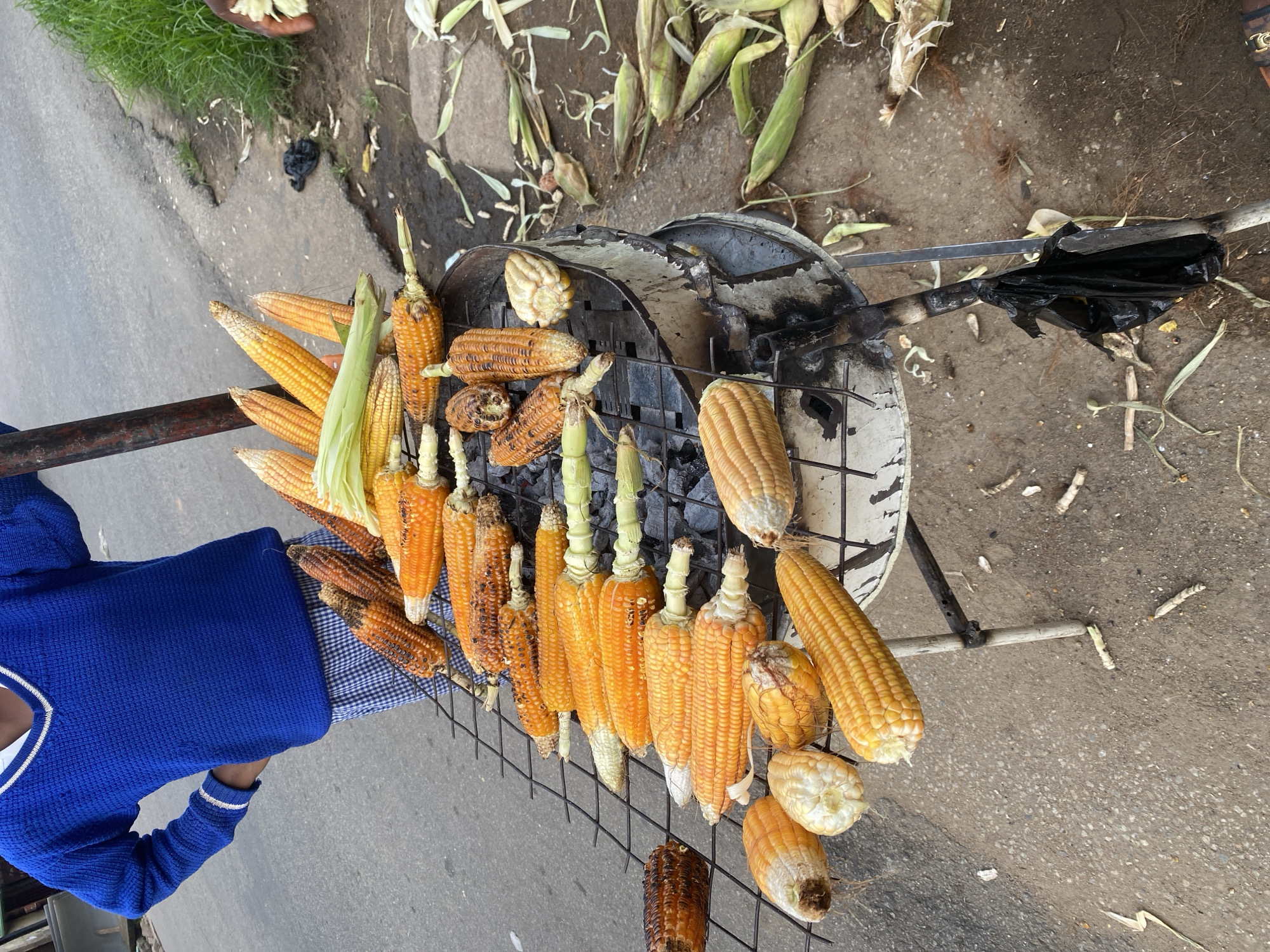 A street vendor in Jos, Nigeria, is roasting corn over a charcoal grill. The corn, some still in their husks, is arranged on a metal rack above the glowing coals. The vendor, wearing a blue sweater, stands beside the grill, ready to serve customers. The scene is set on a paved road with scattered debris and vegetation along the side, suggesting an outdoor market or roadside stall. The time of day appears to be daytime, with natural lighting. The overall impression is one of everyday commerce in a bustling Nigerian city.