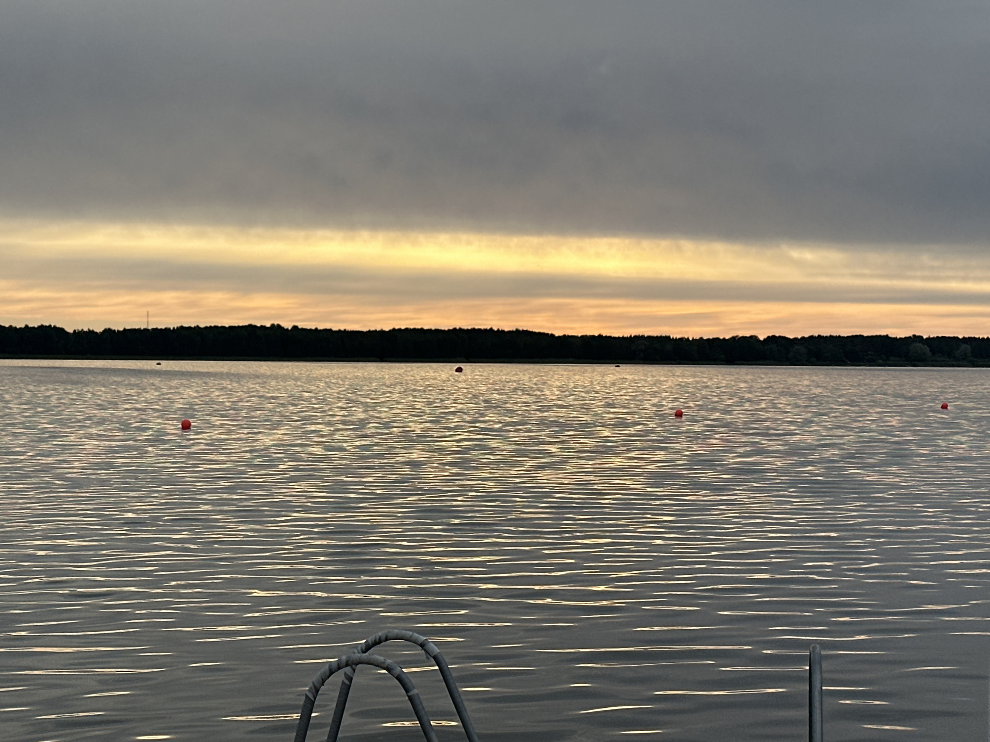 This is a photograph of a lake at sunset with a line of trees in the background. The sky is cloudy with a strip of orange and yellow light near the horizon. There are several red buoys floating on the water. In the foreground, part of a metal ladder is visible, suggesting the photo was taken from a dock or boat. The water is rippled, reflecting the colors of the sky. The location is not identifiable as Tallinn, Estonia.