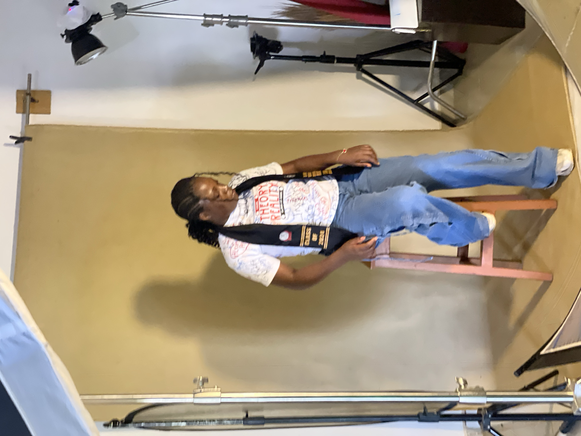A person is lying on a small pink chair against a tan backdrop in a studio setting. They are wearing a white t-shirt with text and graphics, blue jeans, and white socks. A black sash with gold text and emblems is draped over their shoulder. The sash reads "CLASS OF 2020". The person has dark braided hair and appears to be relaxed or in a staged pose.
Visible studio equipment includes lighting stands with lights and a camera stand. The overall scene suggests a professional photoshoot. The lighting appears to be artificial, typical of an indoor studio. No specific time of day or weather conditions can be determined from the image. The location context of Durumi, Nigeria is provided, but no visual cues within the image confirm this specific location. The person's expression is not clearly discernible, but the pose and setting imply a planned activity.