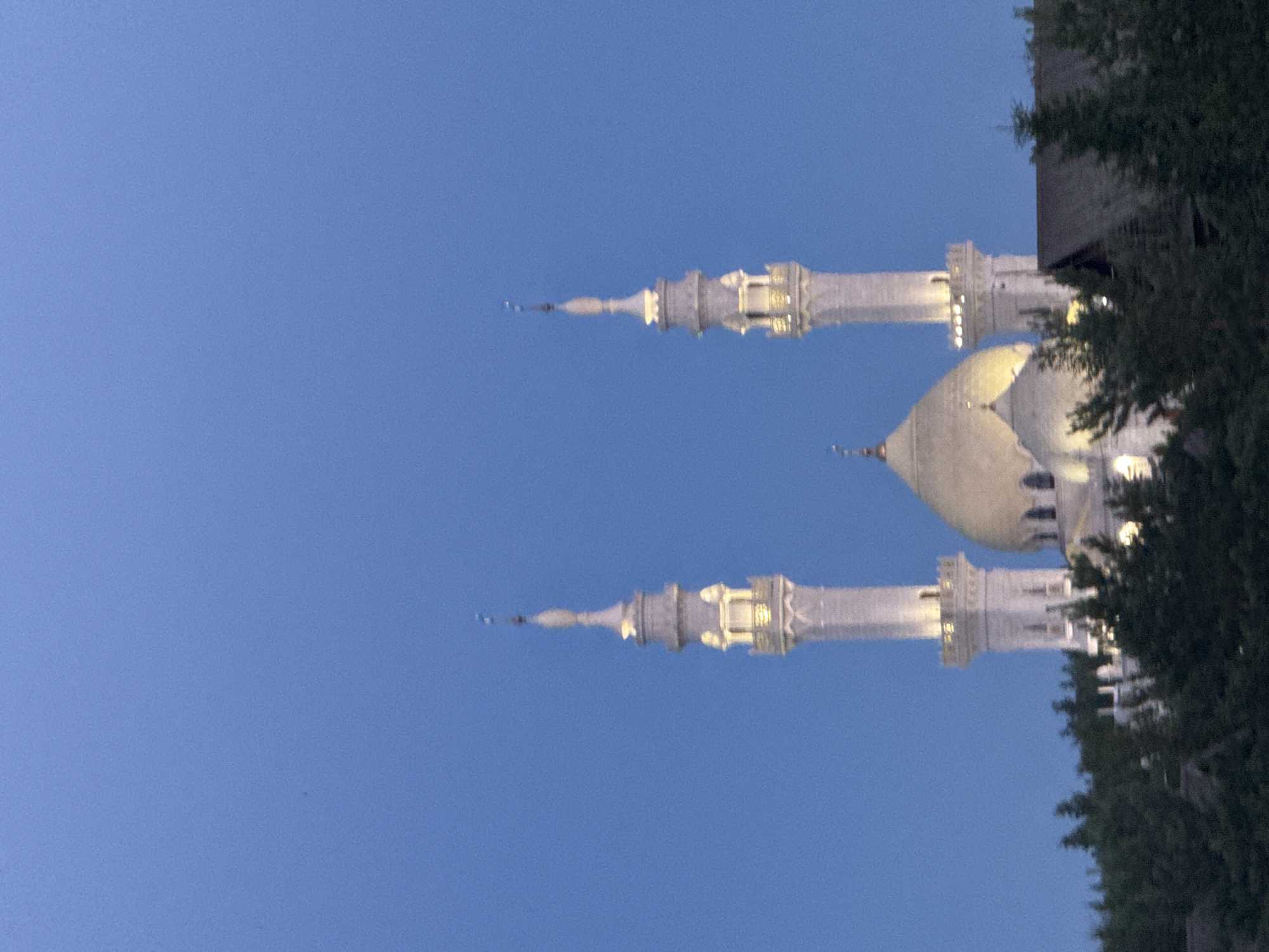 The image captures the White Mosque in Privolzhskiy, Russia, against a twilight blue sky. Two prominent minarets, illuminated from within, flank a central dome. The mosque's white architecture stands out against the darkening sky. In the foreground, a dense line of dark green pine trees partially obscures the lower portion of the mosque and a dark-colored roofline visible on the right. The overall scene is peaceful, with the mosque appearing to be lit up as dusk settles.