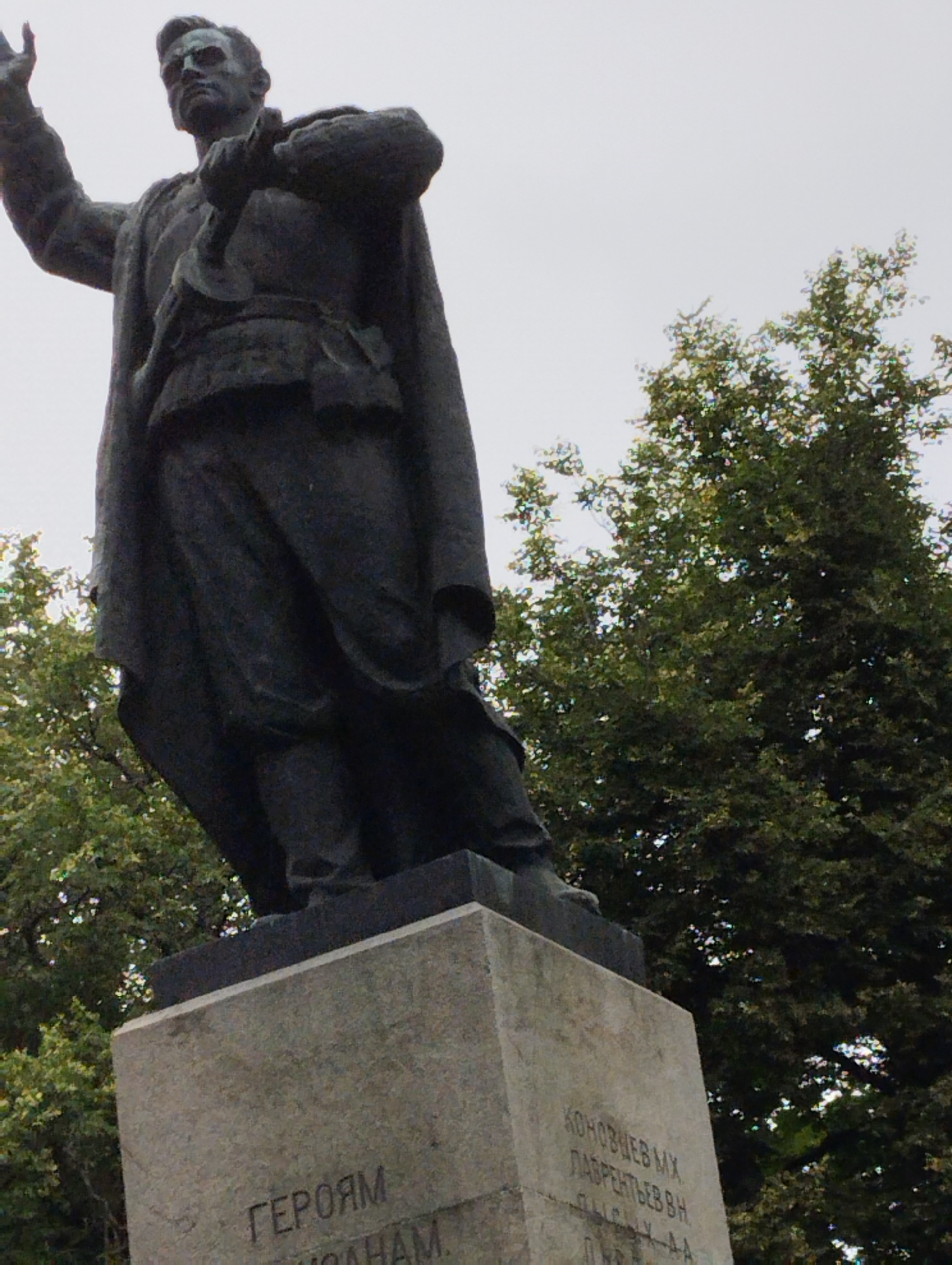 A low-angle, full shot captures a dark, weathered statue of a man in uniform, possibly a soldier or a military leader, standing proudly on a concrete pedestal. The statue is depicted from the waist up, with the figure's right arm raised in a gesture of salute or command, and a hand clutching a baton or rifle. The man wears a coat draped over his shoulders and a belt cinched around his waist. The statue is positioned outdoors, with lush green trees forming a dense backdrop. The sky above is a pale, overcast gray, suggesting a cloudy day.
Visible text on the pedestal reads in Russian: "ГЕРОЯМ" (To the Heroes) followed by what appears to be names and possibly dates, though some are obscured or difficult to discern. Further down, another line reads "КОНОНОВУ МХ" (Kononov M.Kh.) and "ЛАВРЕНТЬЕВУ В.Н." (Lavrentyev V.N.). This monument is located in Kingisepp, Russia. The overall mood is solemn and commemorative, honoring significant figures from history.