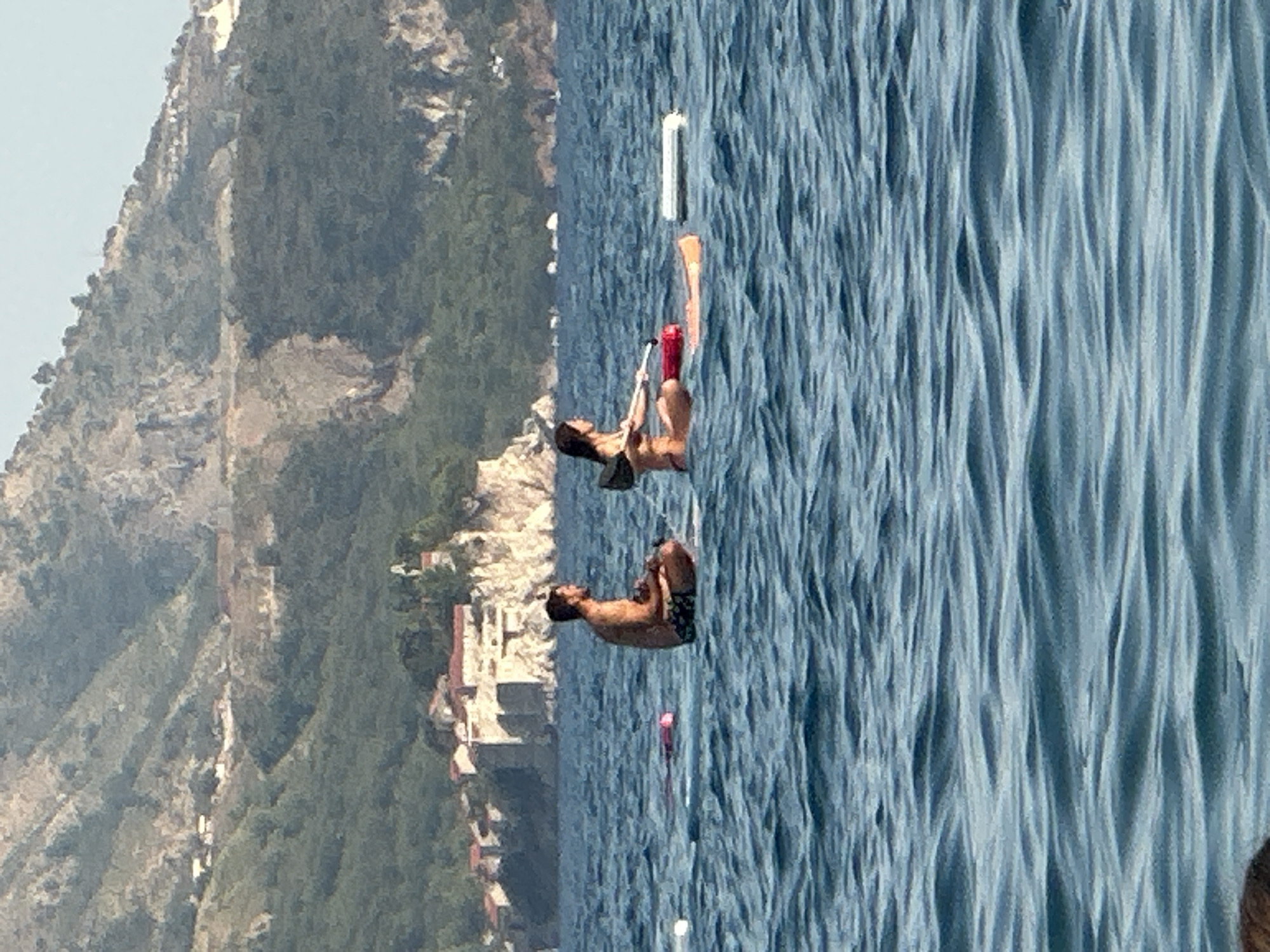 In the city of Boreti, Montenegro, two individuals are seen on paddleboards in the sea. The person on the left, a male, is seated cross-legged on a paddleboard, facing right. The person on the right, a female, is seated on a paddleboard as well, holding a paddle and facing left. Both individuals are wearing swimwear. The sea is a deep blue with visible waves, and a mountainous, tree-covered landscape with buildings is visible in the background. Several buoys and other floating objects are also present in the water.
