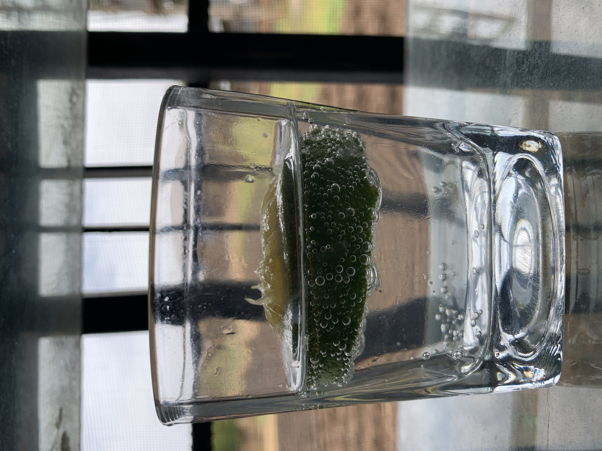 A clear, square glass is filled with water and contains a slice of lime. Bubbles are clinging to the lime and suspended in the water. The glass sits on a textured surface, likely a table, which appears to be indoors near a window with blinds or a screen. The location is unlikely to be Rafin Par, Nigeria, based on the setting depicted in the image.