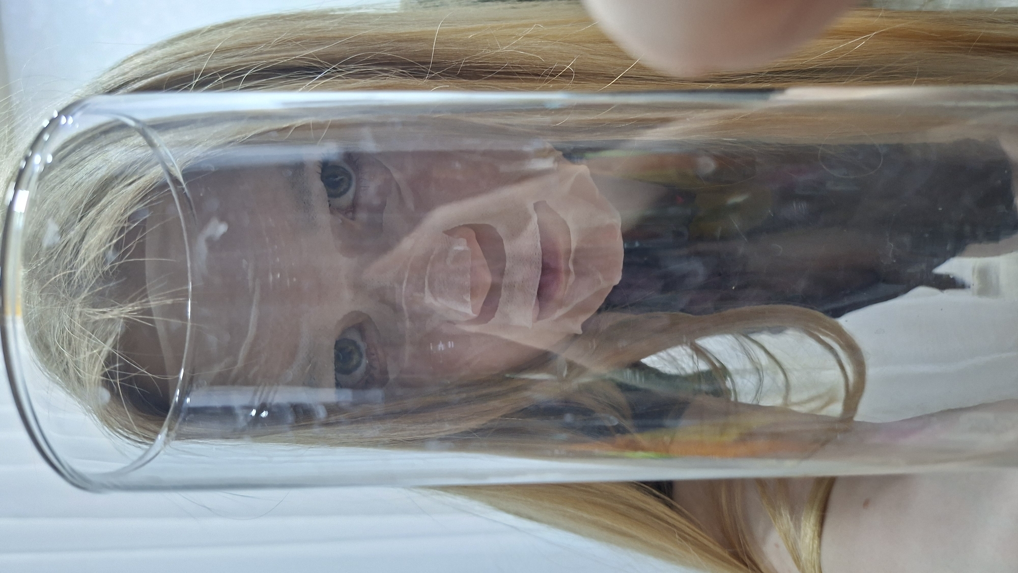 A woman with blonde hair is seen through a transparent tube. She has fair skin and light-colored eyes. She wears a facial mask. The image appears to be taken in Chaikovsky, Russia, based on the prompt.
