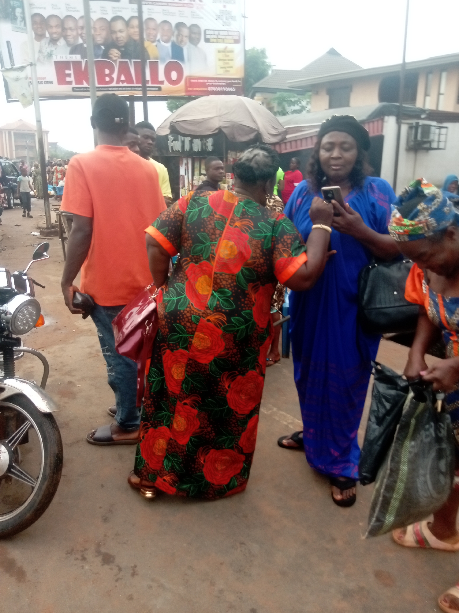 The image captures a street scene in Nkpor, Nigeria. Three women are standing in the foreground, two of whom are in focus. The woman in the center is wearing a blue dress and using a smartphone. To her left, a woman wears a dark dress with red and orange floral patterns. To the right, a third woman is partially visible, wearing a blue and orange patterned top. Behind them is a man in an orange shirt and jeans. A parked motorcycle is partially visible on the left. In the background, a crowd of people is milling about. A banner or poster is displayed overhead, and a food vendor tent with an umbrella roof is in the middle ground. The buildings in the background appear to be a mix of residential and commercial structures. The ground is paved.