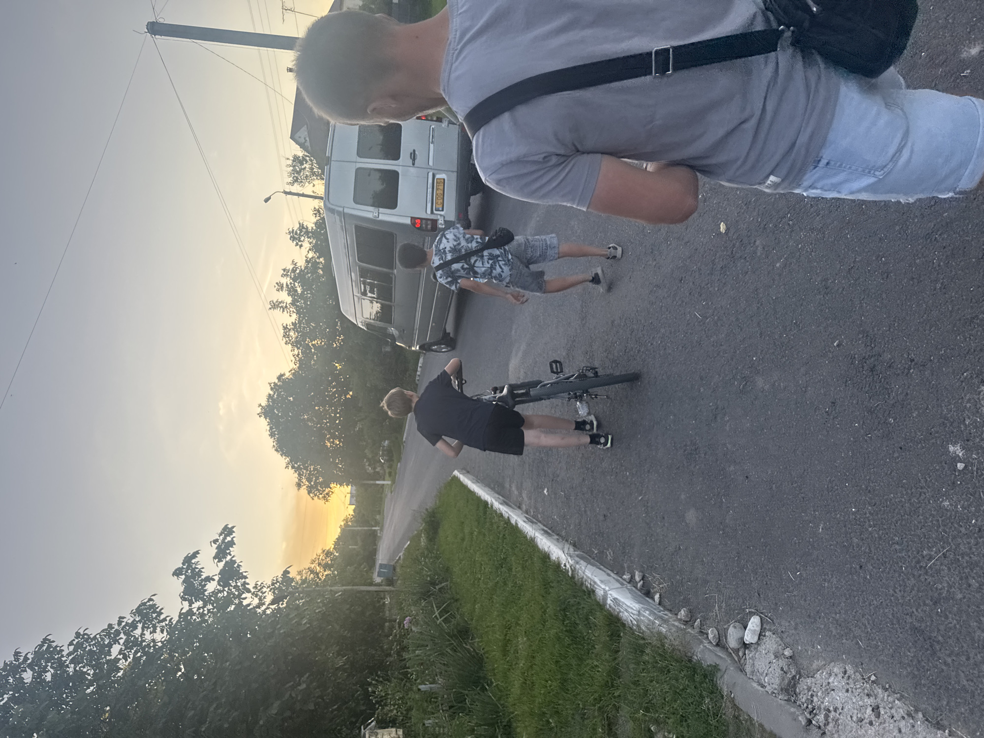 This media file depicts an outdoor scene with a group of people and a vehicle. In the foreground, a person in a grey t-shirt and denim shorts is standing, facing away from the camera. Behind them, a white van is parked on a paved road. Two other individuals are visible near the van. One, wearing a black t-shirt and shorts, is standing beside a bicycle, which is leaning against them. The other person is wearing a patterned shirt and shorts and is walking away from the van.
The scene appears to be set during the late afternoon or early evening, suggested by the warm, soft lighting and the orange glow on the horizon. The sky is a pale grey, transitioning to yellow and orange near the horizon, indicating sunset or sunrise. Trees and foliage line the sides of the road, suggesting a suburban or rural setting. The road surface is asphalt, with a concrete curb and grass bordering it. A street light is visible in the background.
No specific event or activity is clearly depicted, other than people being present in the vicinity of a van and a bicycle. The overall mood seems casual and relaxed. There is some visible text on the license plate of the van, which reads "CGG 715". The image is taken from a slightly elevated perspective, looking down and across the scene.